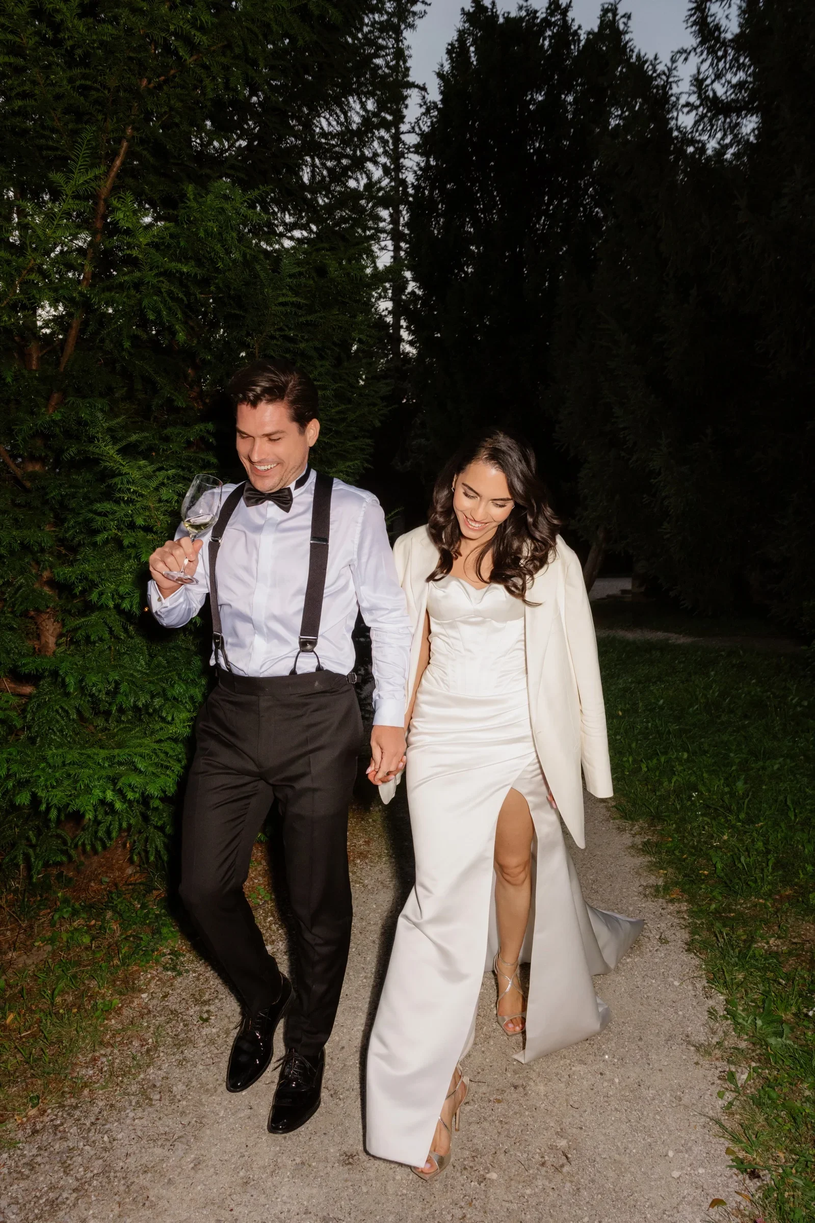 Couple walking hand in hand along a gravel path in the evening, the groom in suspenders and bow tie