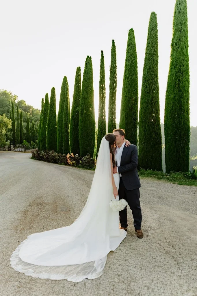Couple kissing on a cypress-lined avenue, long veil trailing behind
