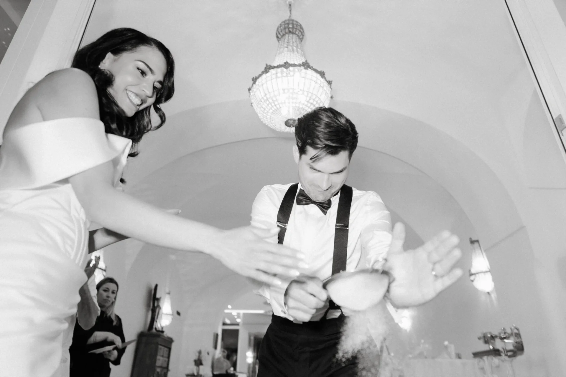 Bride laughing while the groom in suspenders performs a trick under a vaulted ceiling, black and white