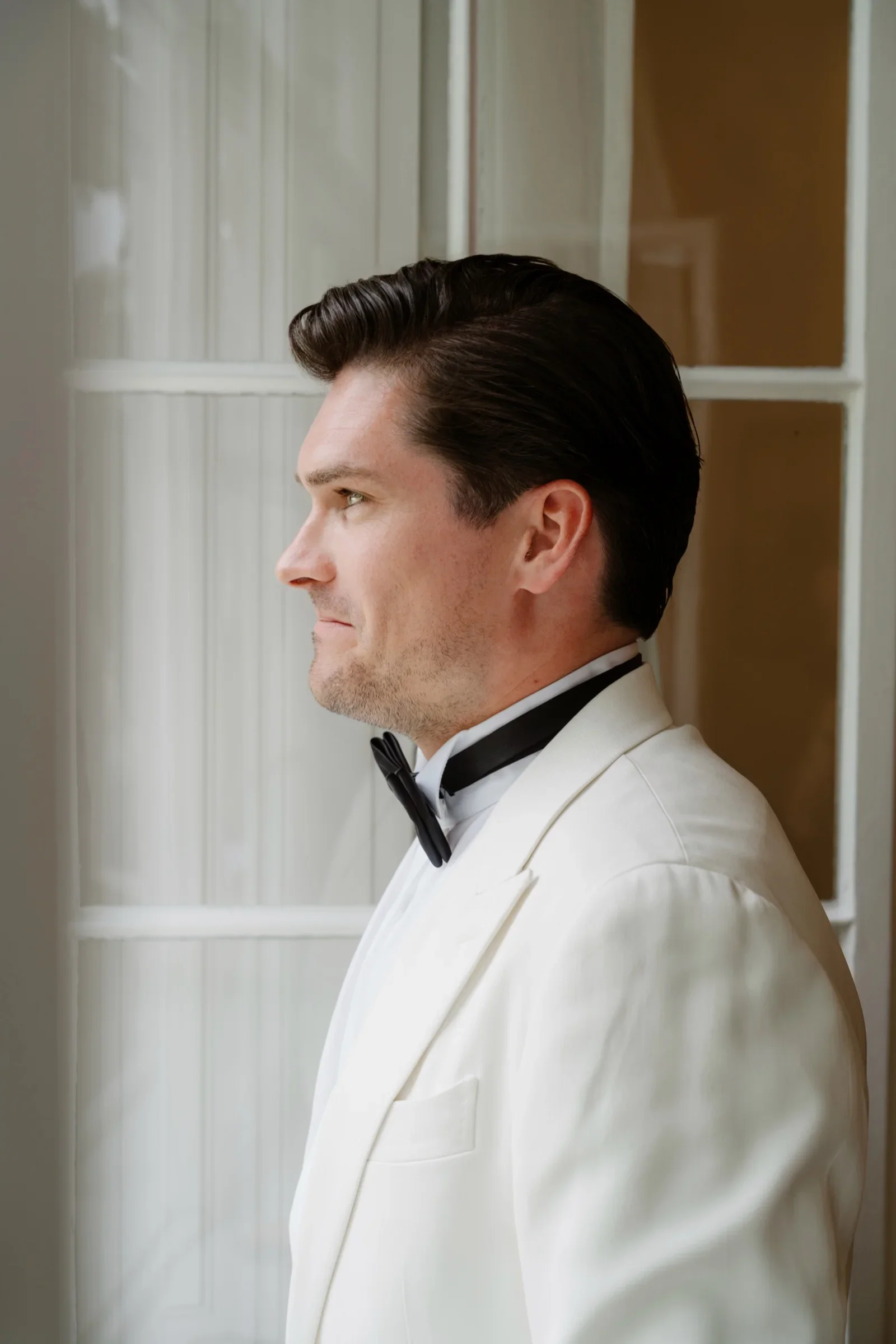 Groom in a white dinner jacket and bow tie looking out a tall window