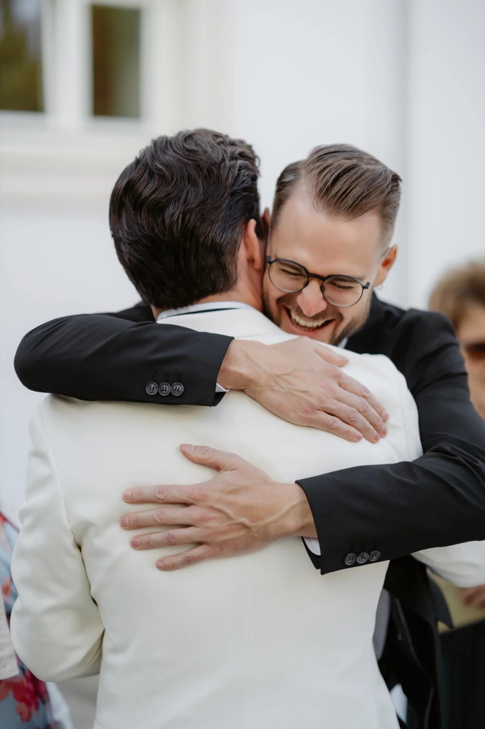 Groom in a white jacket hugging a smiling guest with glasses