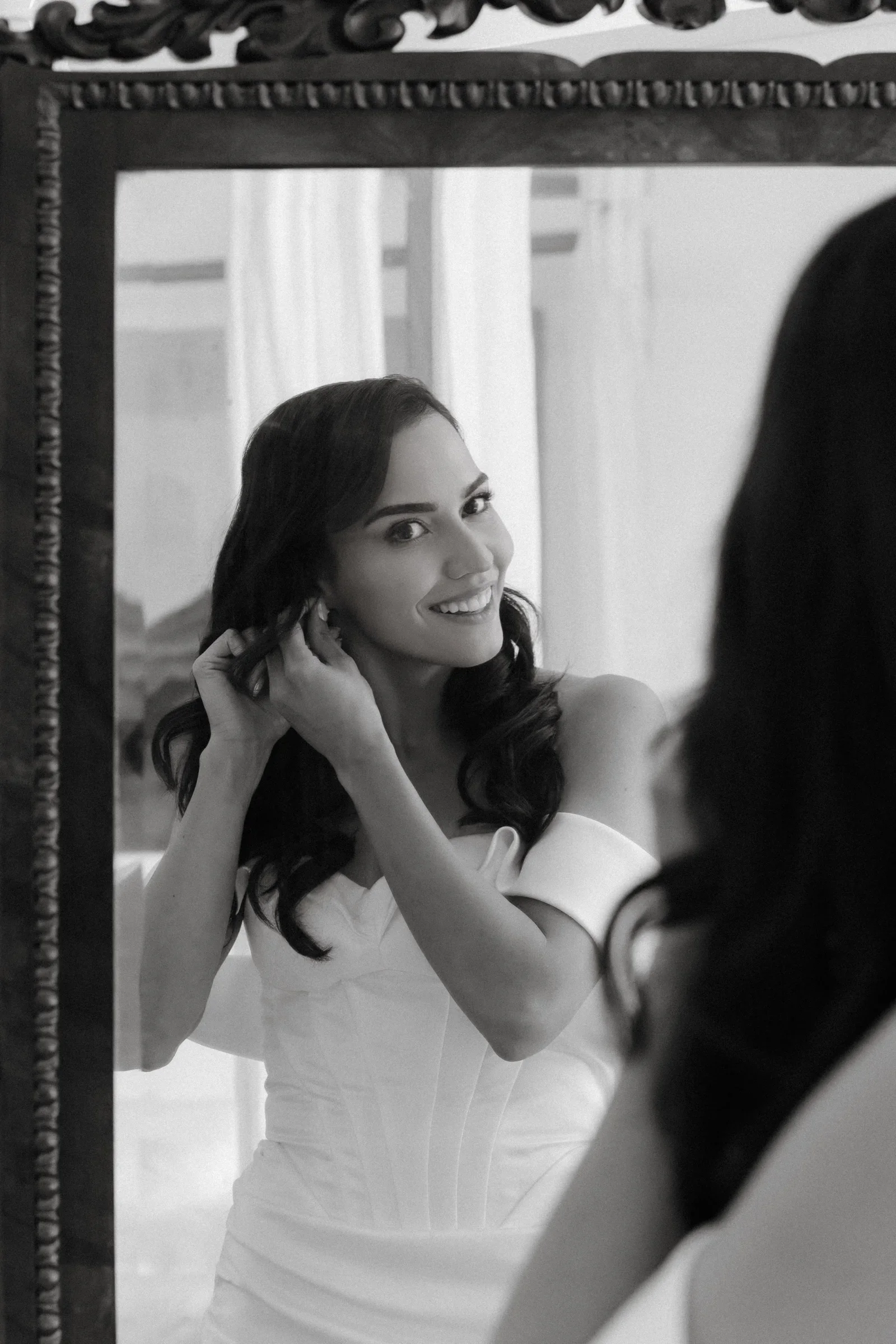 Black and white photo of the bride smiling in a mirror while adjusting her earring