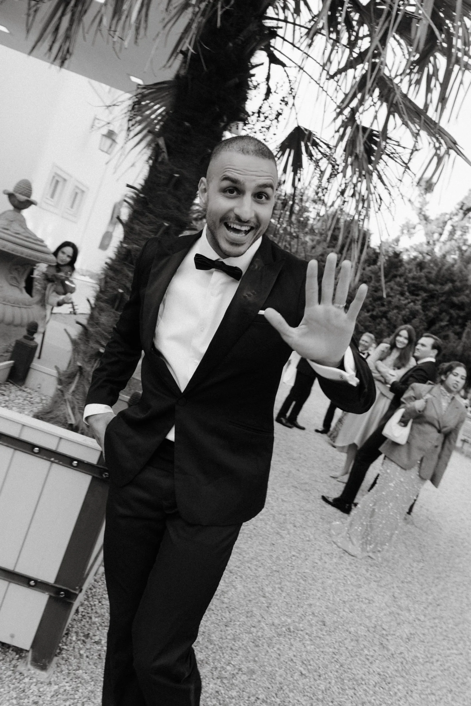 Groom in a black tuxedo waving at the camera on a gravel path, black and white