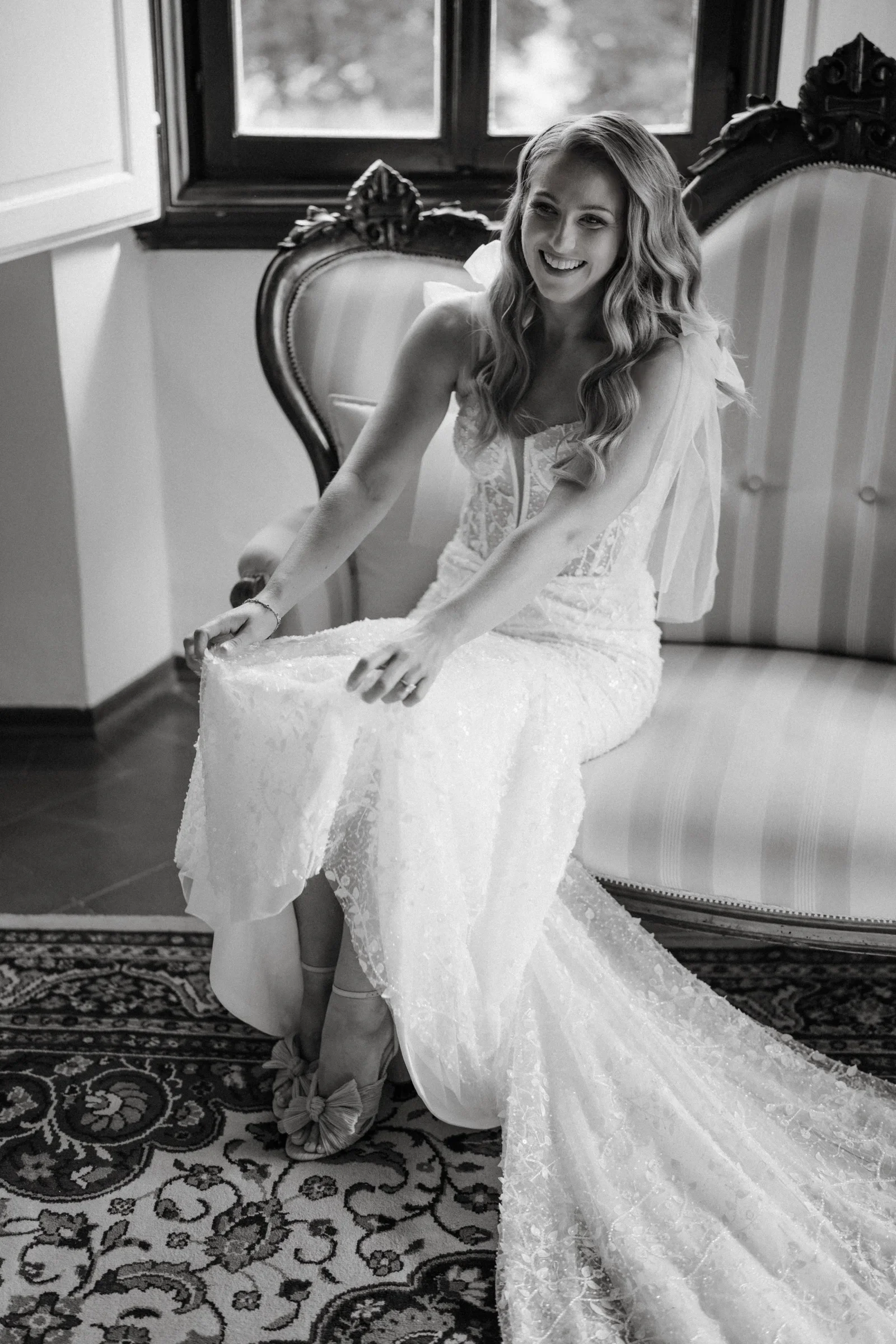 Black and white: the bride seated on a carved chair, lifting the hem of her lace gown