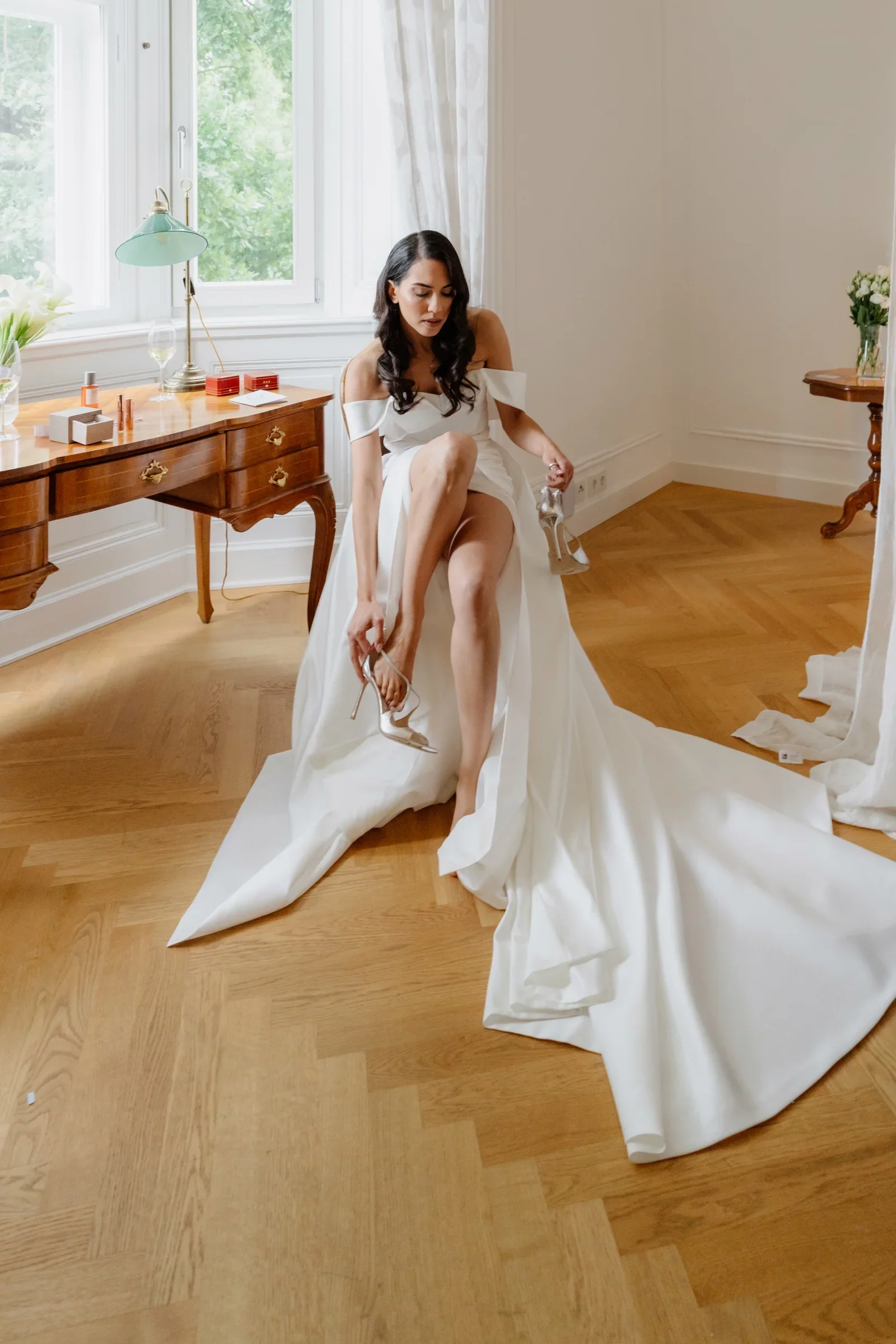 Bride in a wedding gown with a long train sitting by a window putting on a shoe