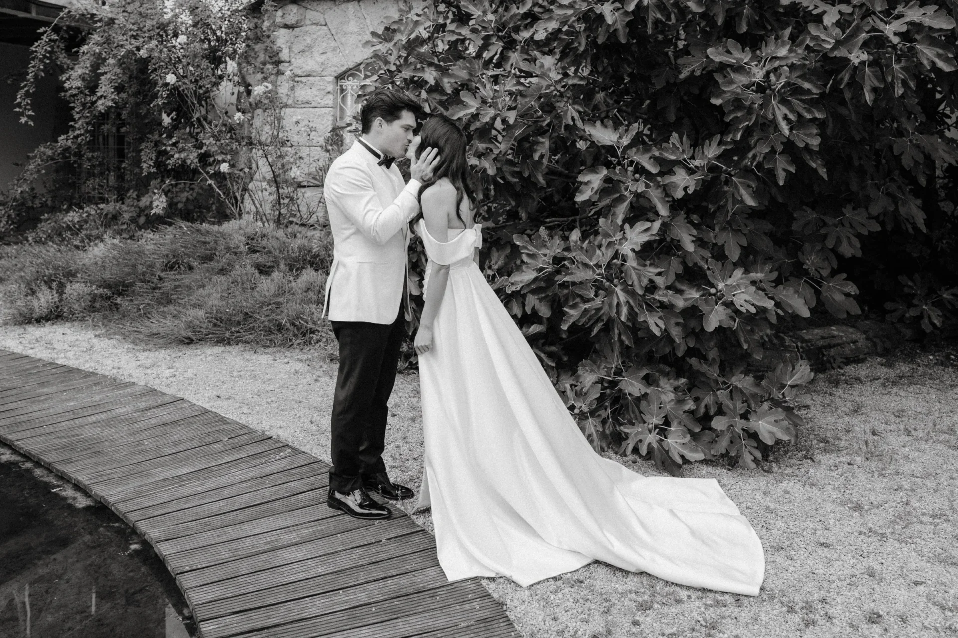 Black and white photo of the couple embracing on a garden path, the bride's long train trailing