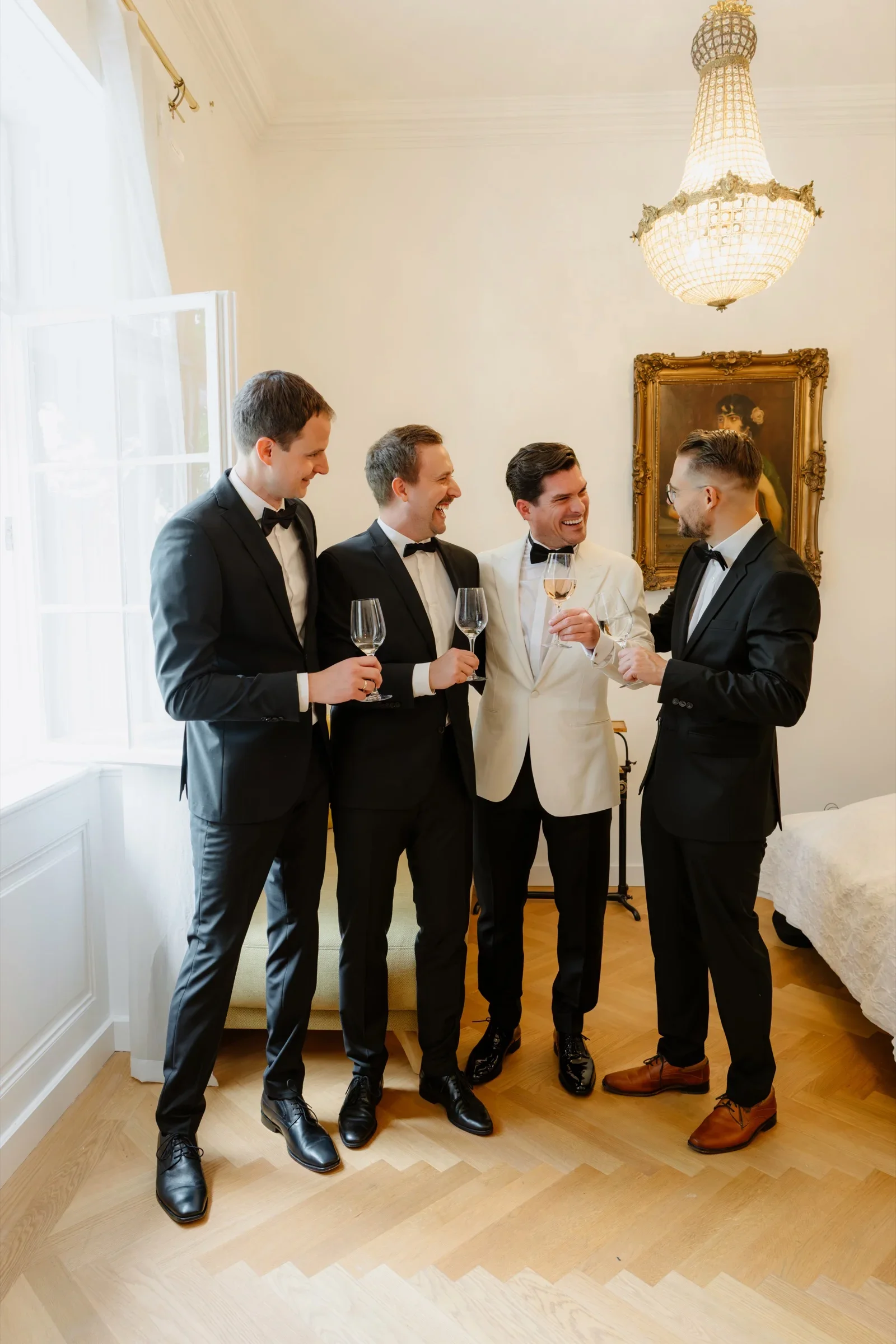 Groom and three groomsmen in tuxedos laughing with glasses of wine under a chandelier