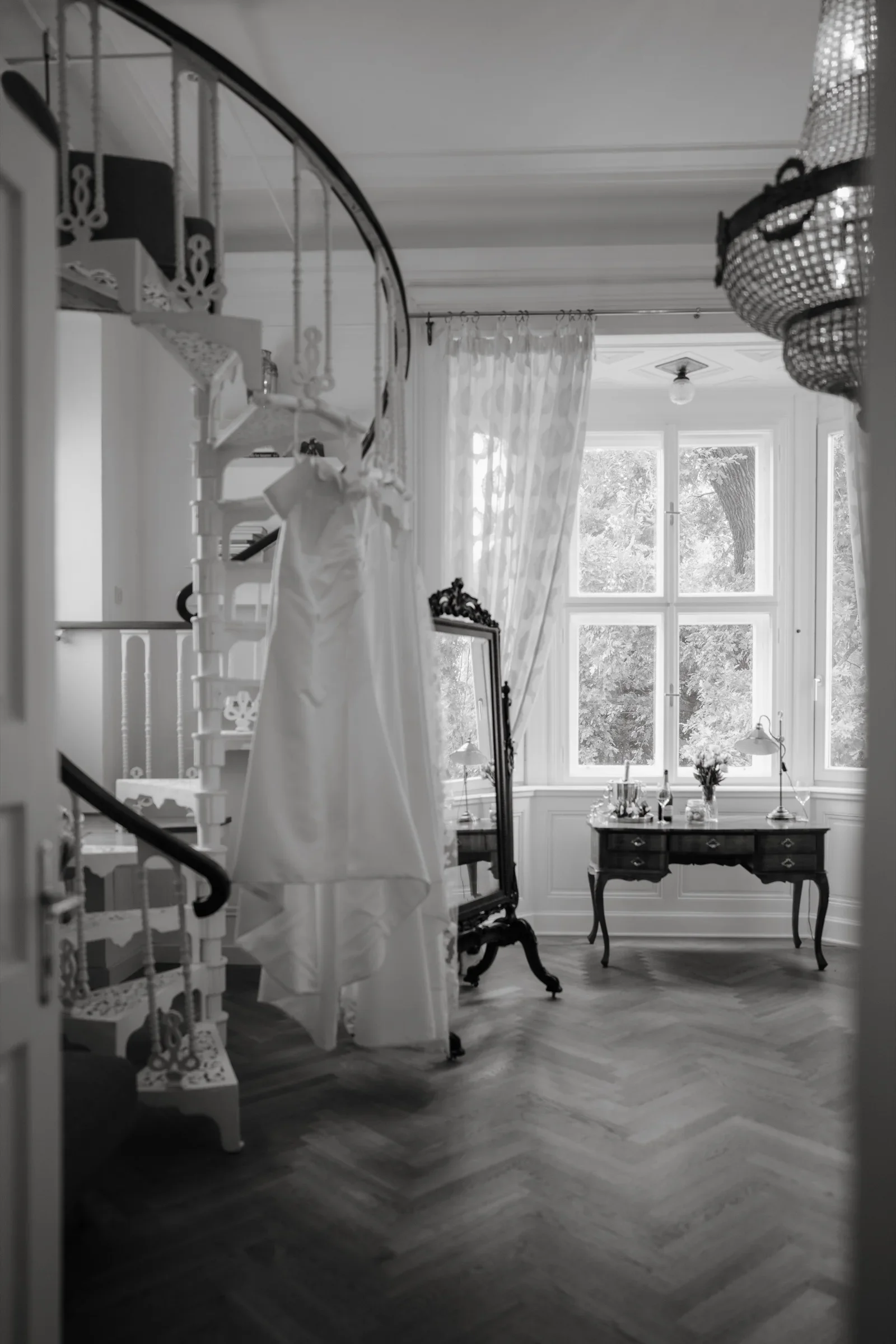 Black and white photo of a wedding dress hanging by a spiral staircase in a bright room