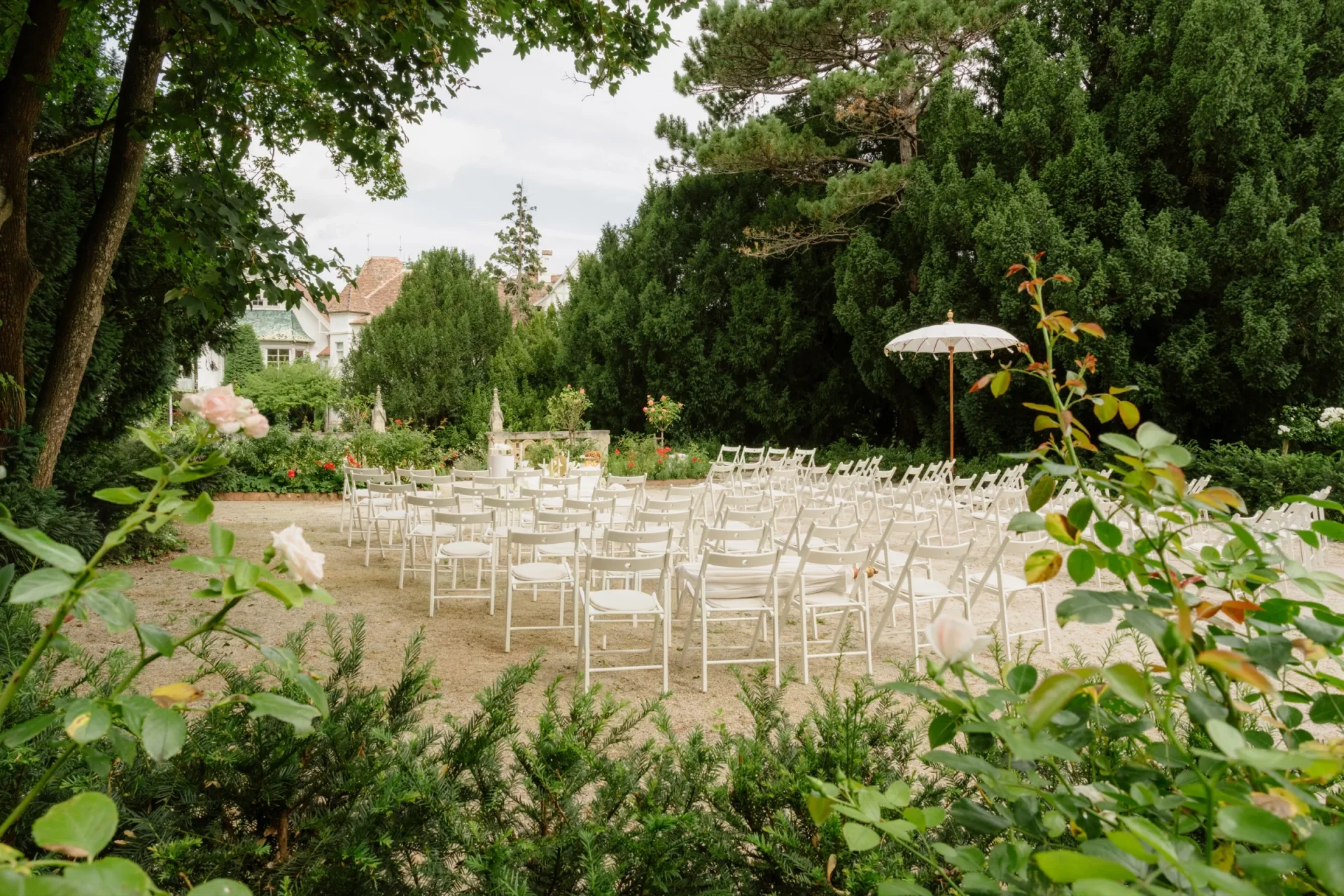White chairs arranged for an outdoor ceremony among green trees and roses
