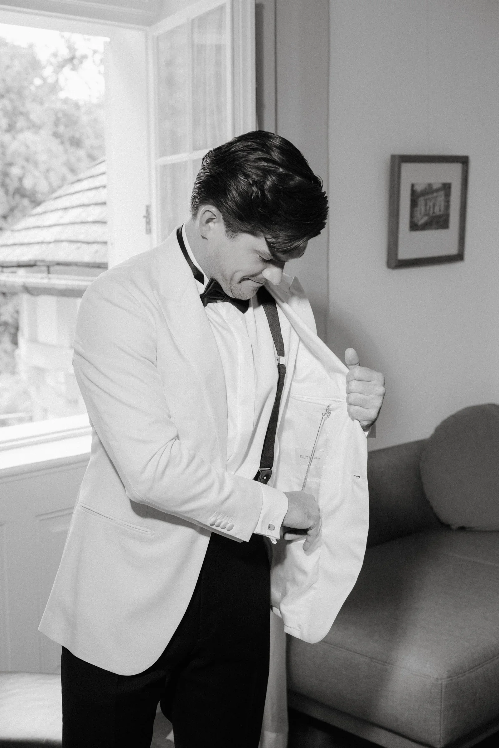 Black and white photo of the groom in a white dinner jacket putting on his jacket beside a window