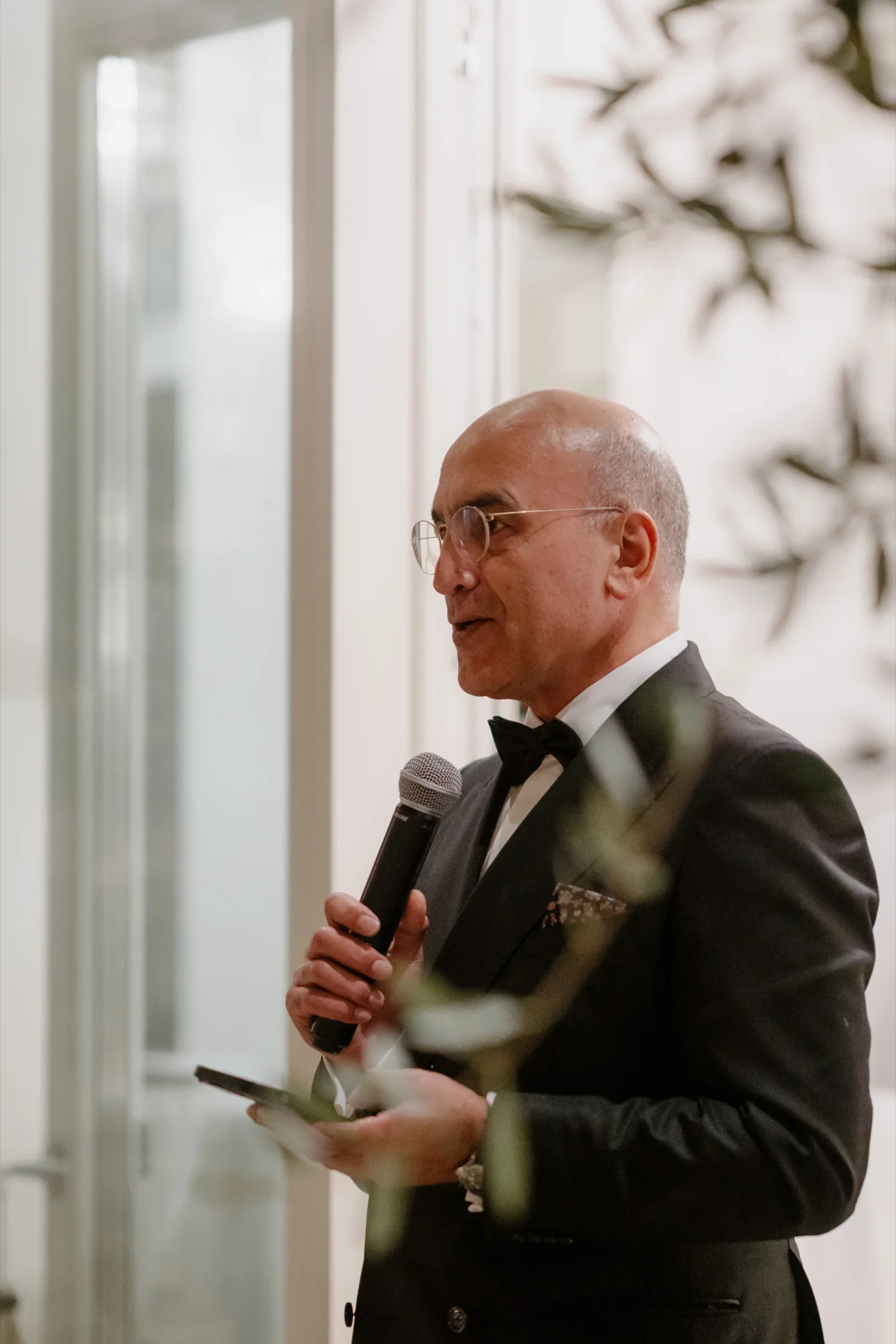 Man in a tuxedo giving a speech into a microphone by a tall window
