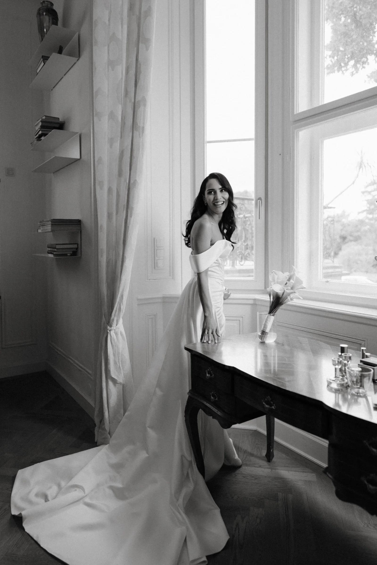 Black and white photo of the bride with a long veil standing by a tall window
