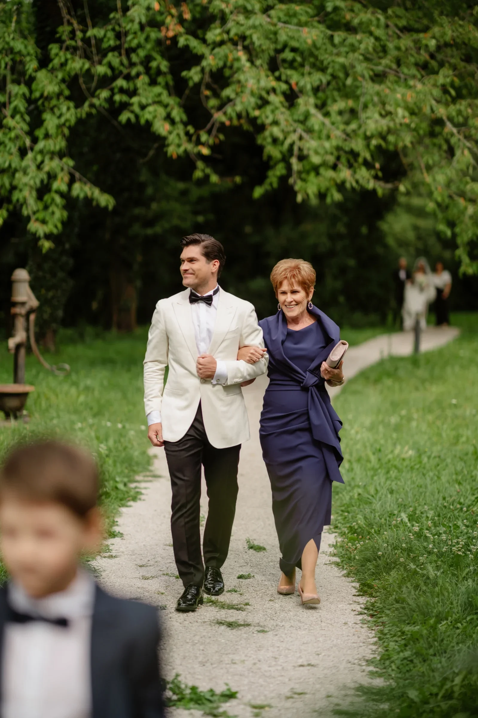 Groom in a white dinner jacket walking with a woman in a purple dress on a garden path