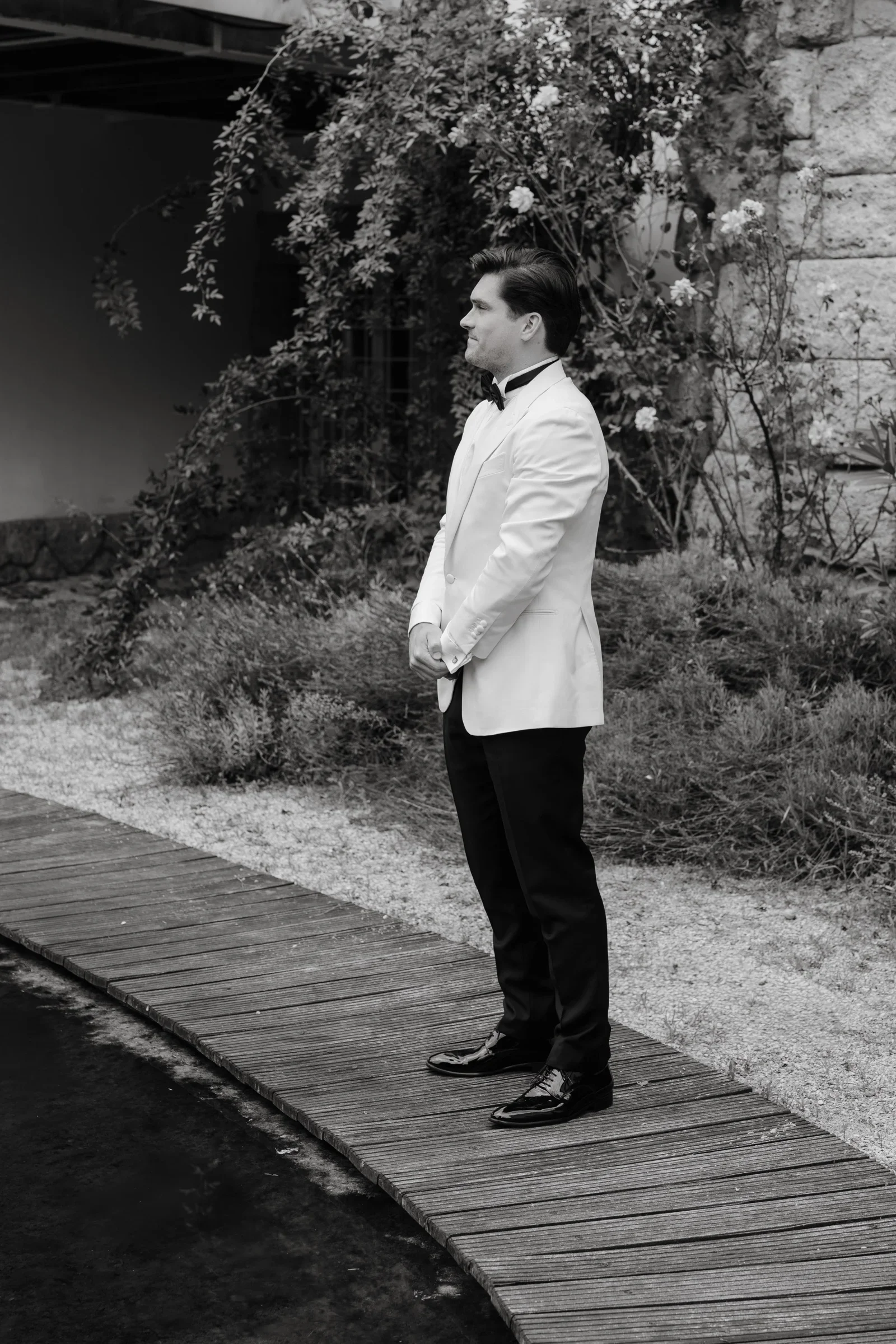 Groom in a white dinner jacket standing on a wooden garden path with his back turned