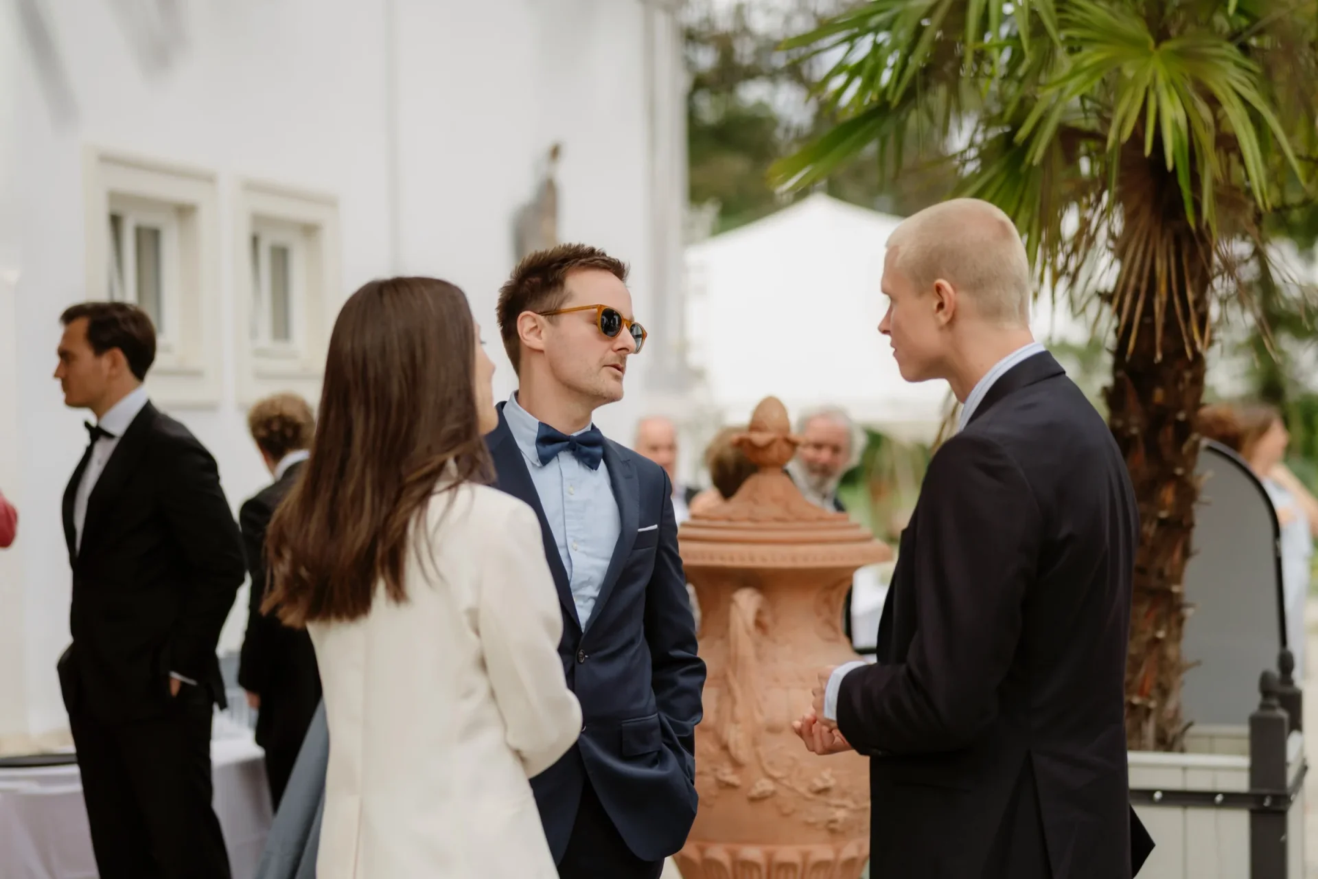 Guests chatting in front of a white villa, one man in sunglasses and a navy suit