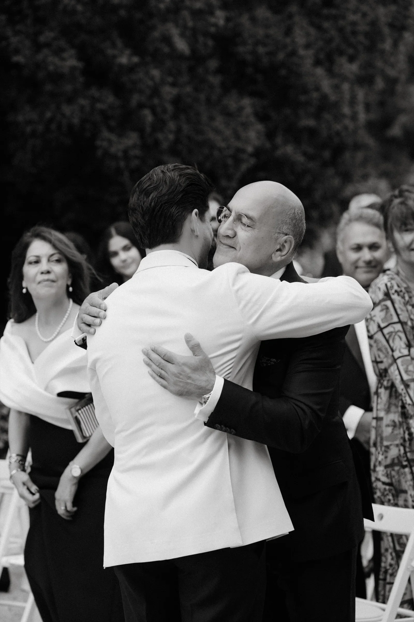 Black and white photo of the groom in a white jacket hugging an older man
