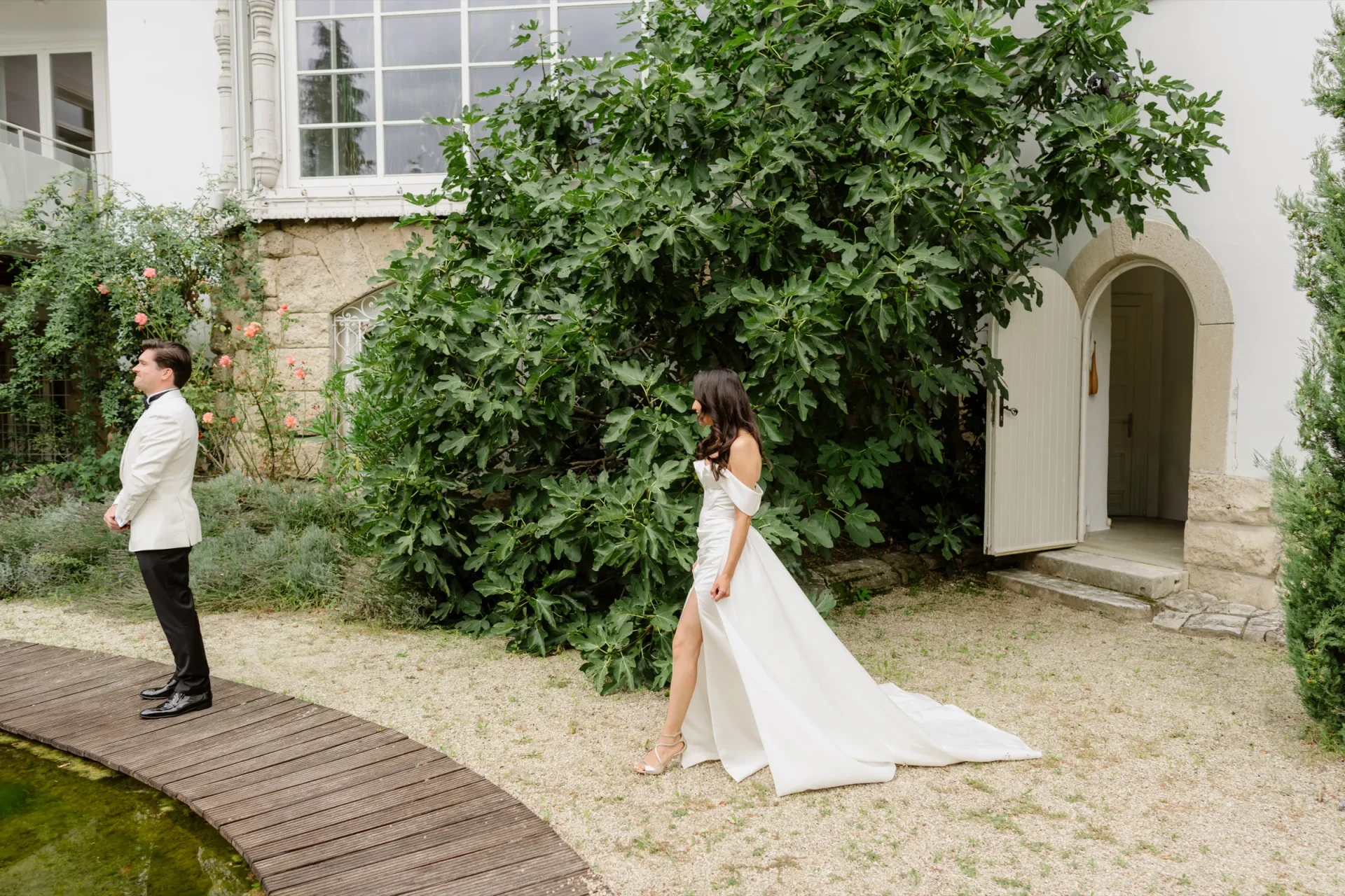 Bride in a gown with a long train walking toward the groom in a sunlit castle garden