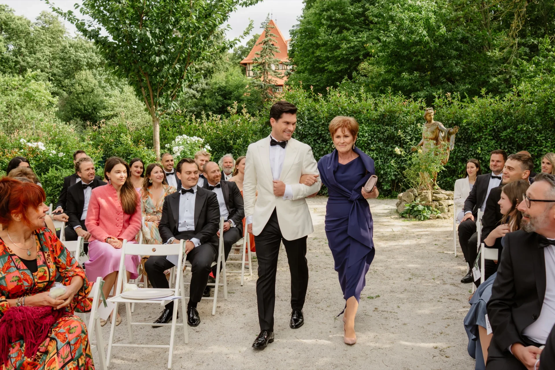 Groom in a white jacket walking with a woman in a blue dress past seated guests