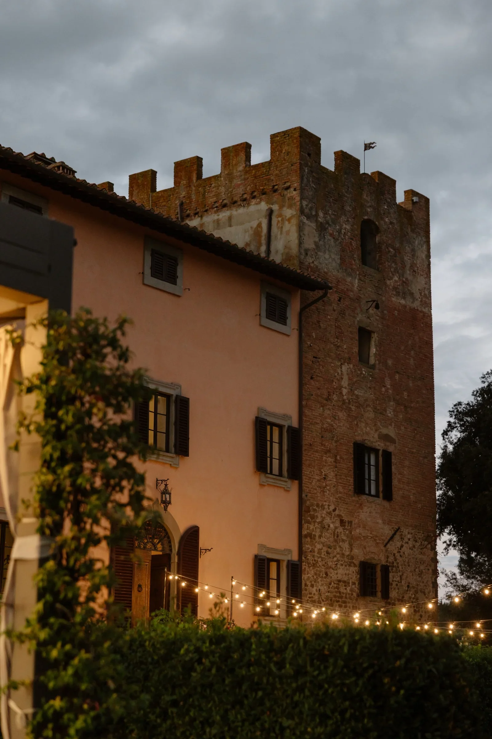 The villa's stone tower lit from below at dusk, festoon lights strung across the garden