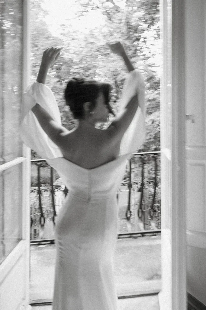 Bride throwing open balcony doors, arms raised, seen from behind