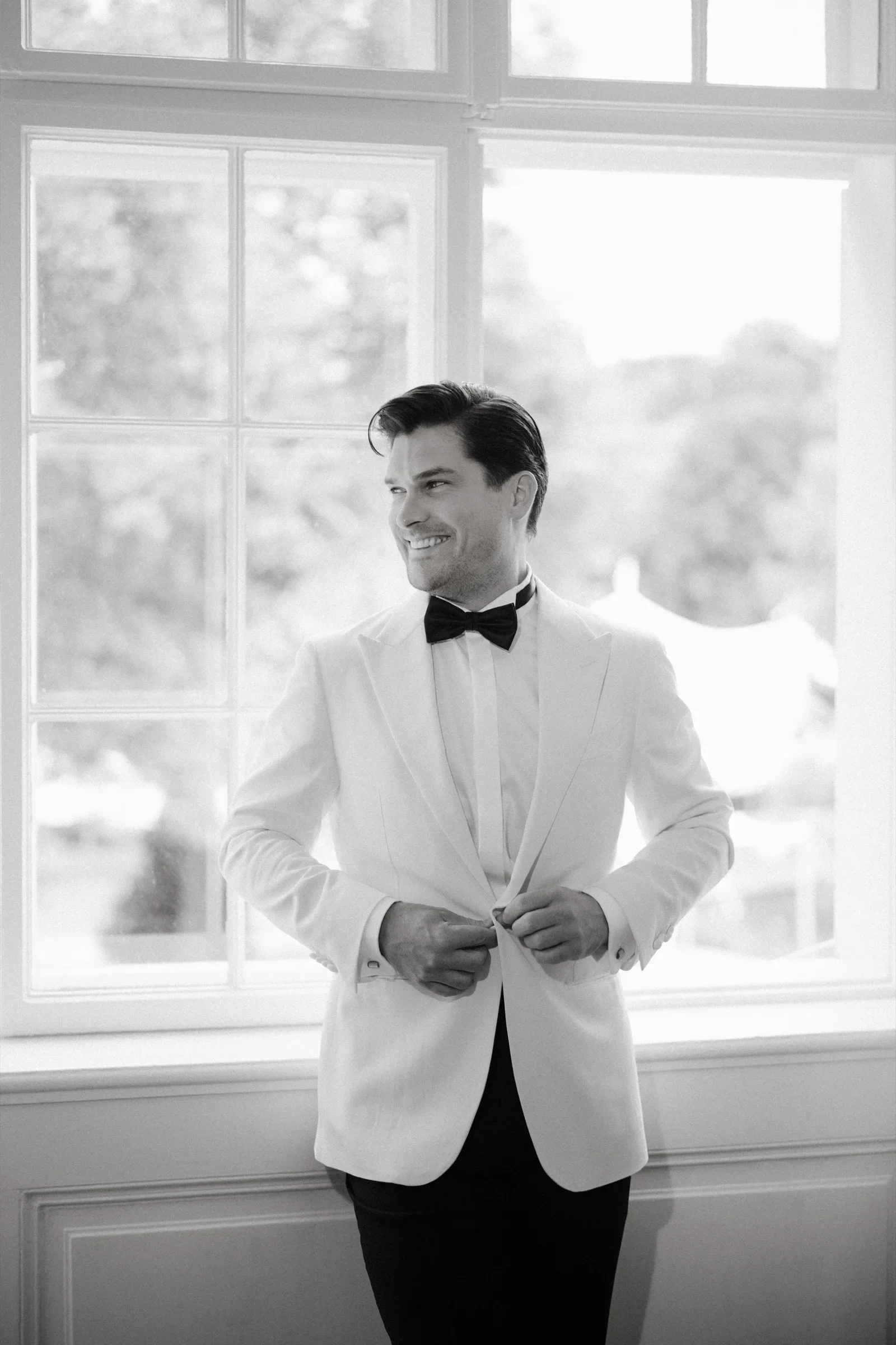 Black and white photo of the groom buttoning his white dinner jacket and smiling by a window