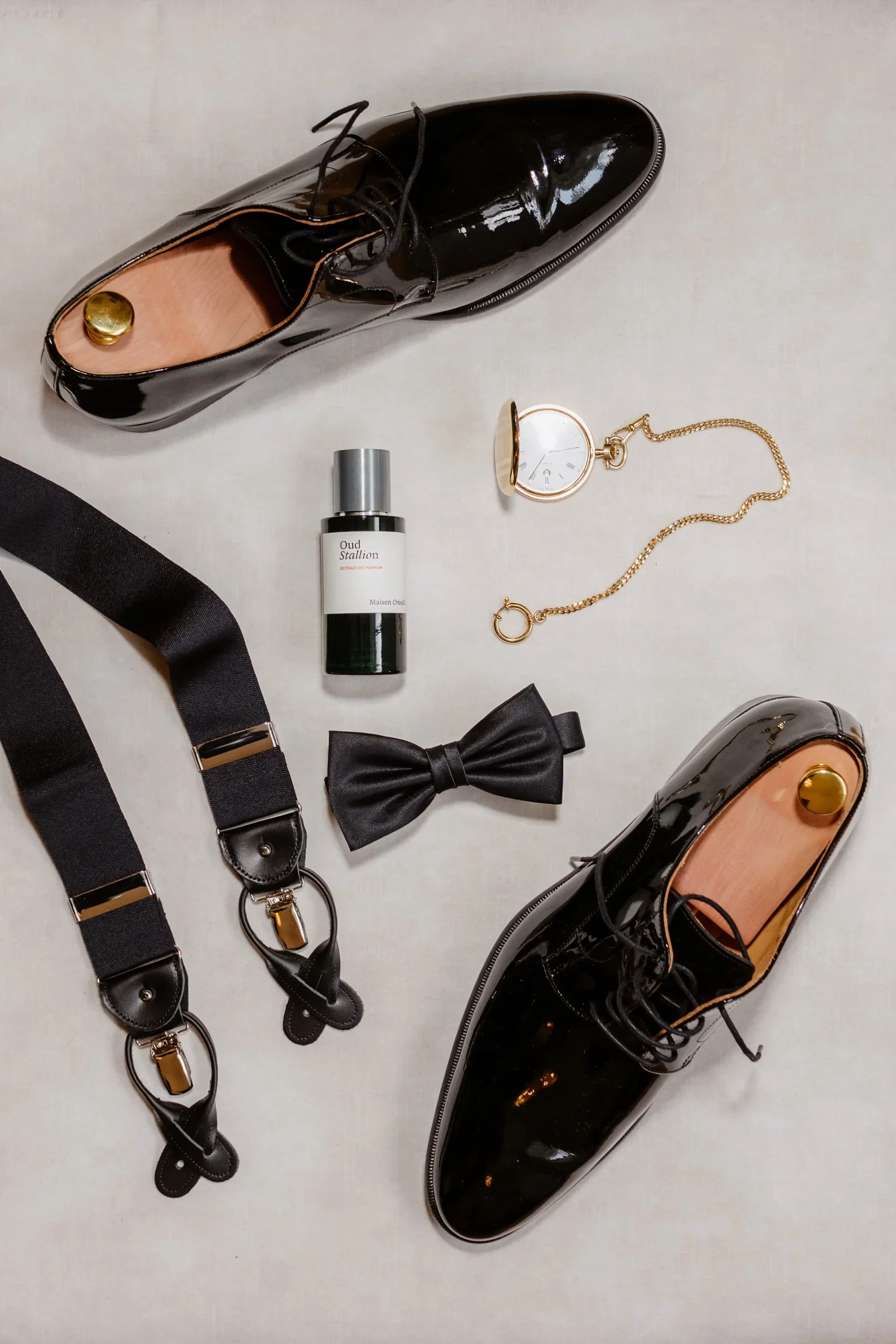 Flat lay of the groom's patent shoes, braces, bow tie, pocket watch and cologne