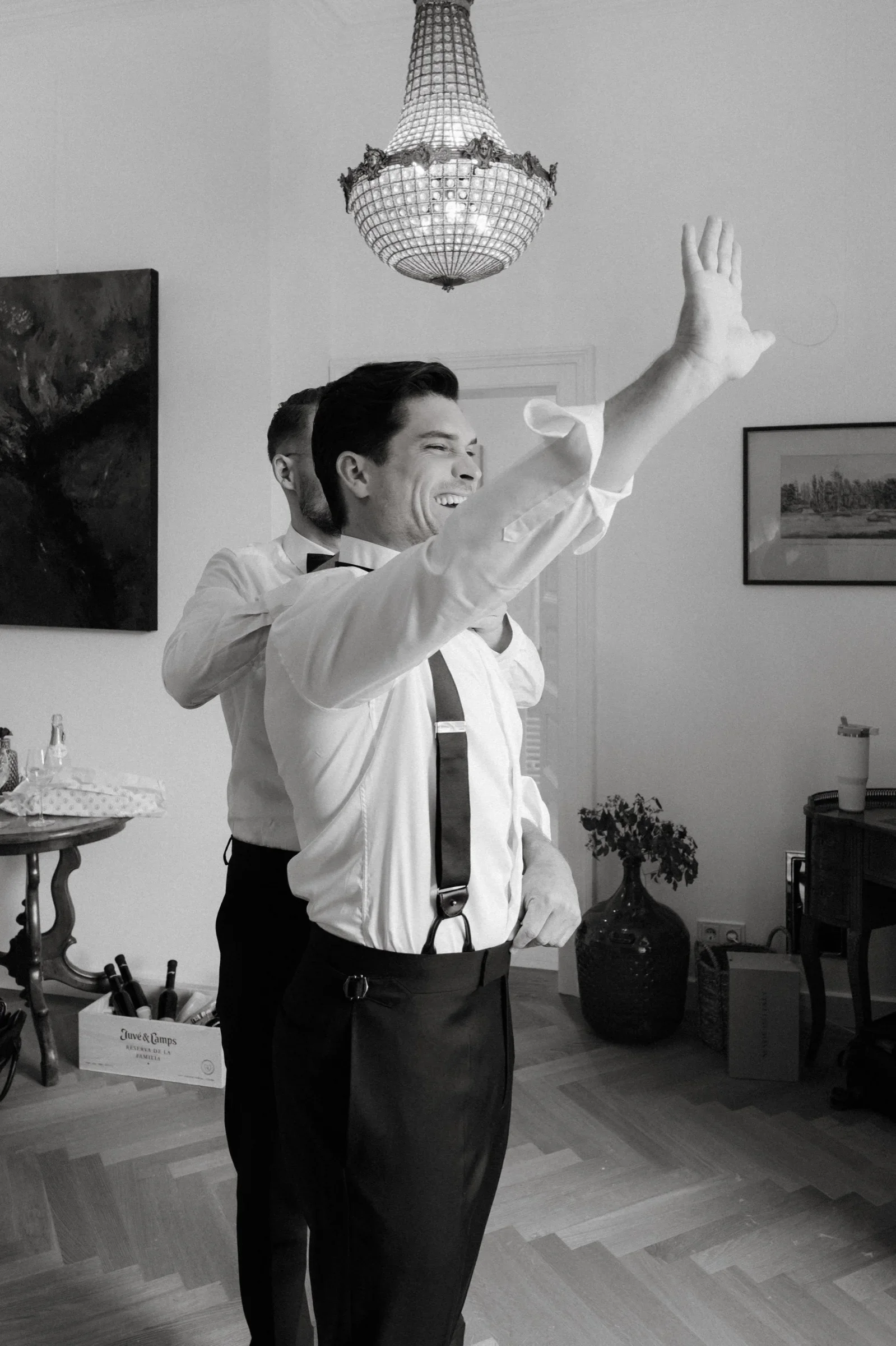 Black and white photo of a groomsman cheering with a raised arm while another helps him dress