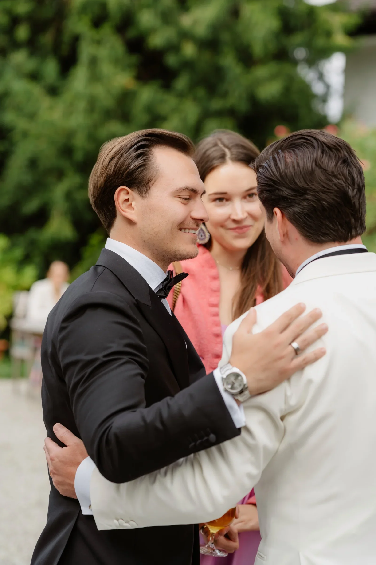 Groom in a black suit embracing a guest while a woman in pink smiles