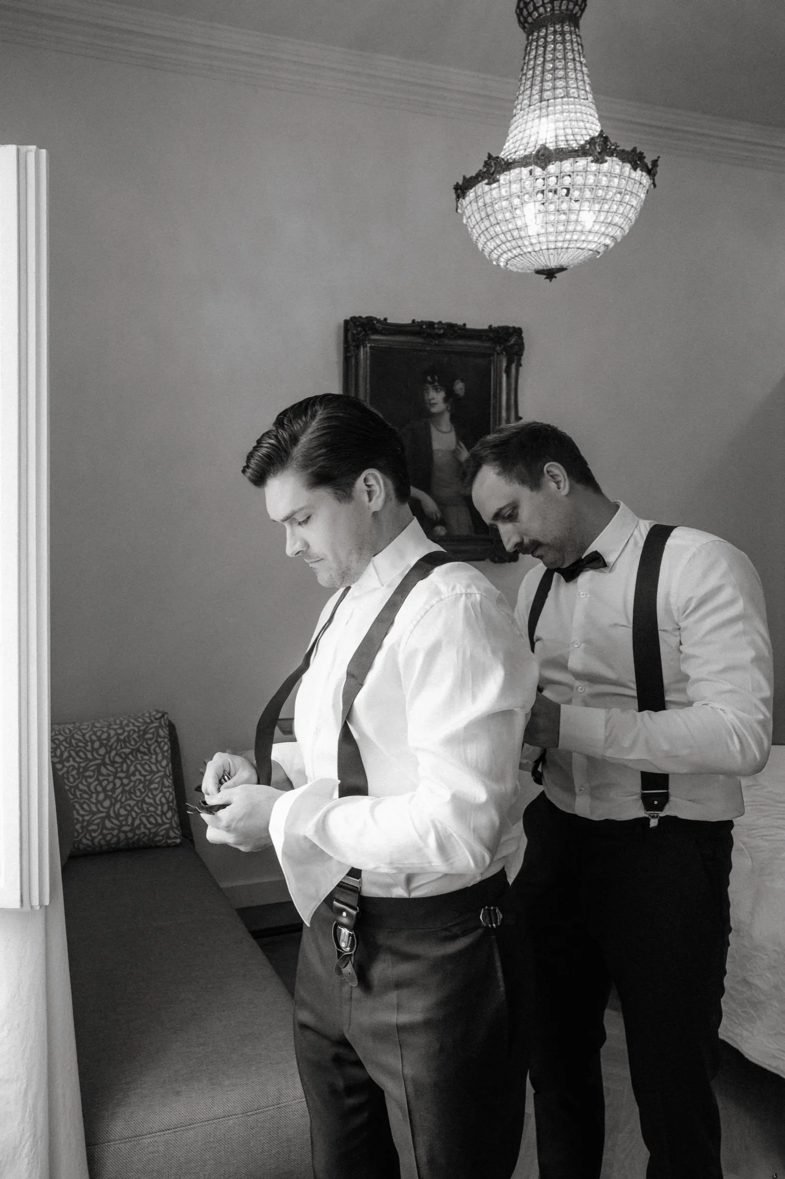 Black and white photo of two groomsmen helping each other with their suspenders under a chandelier