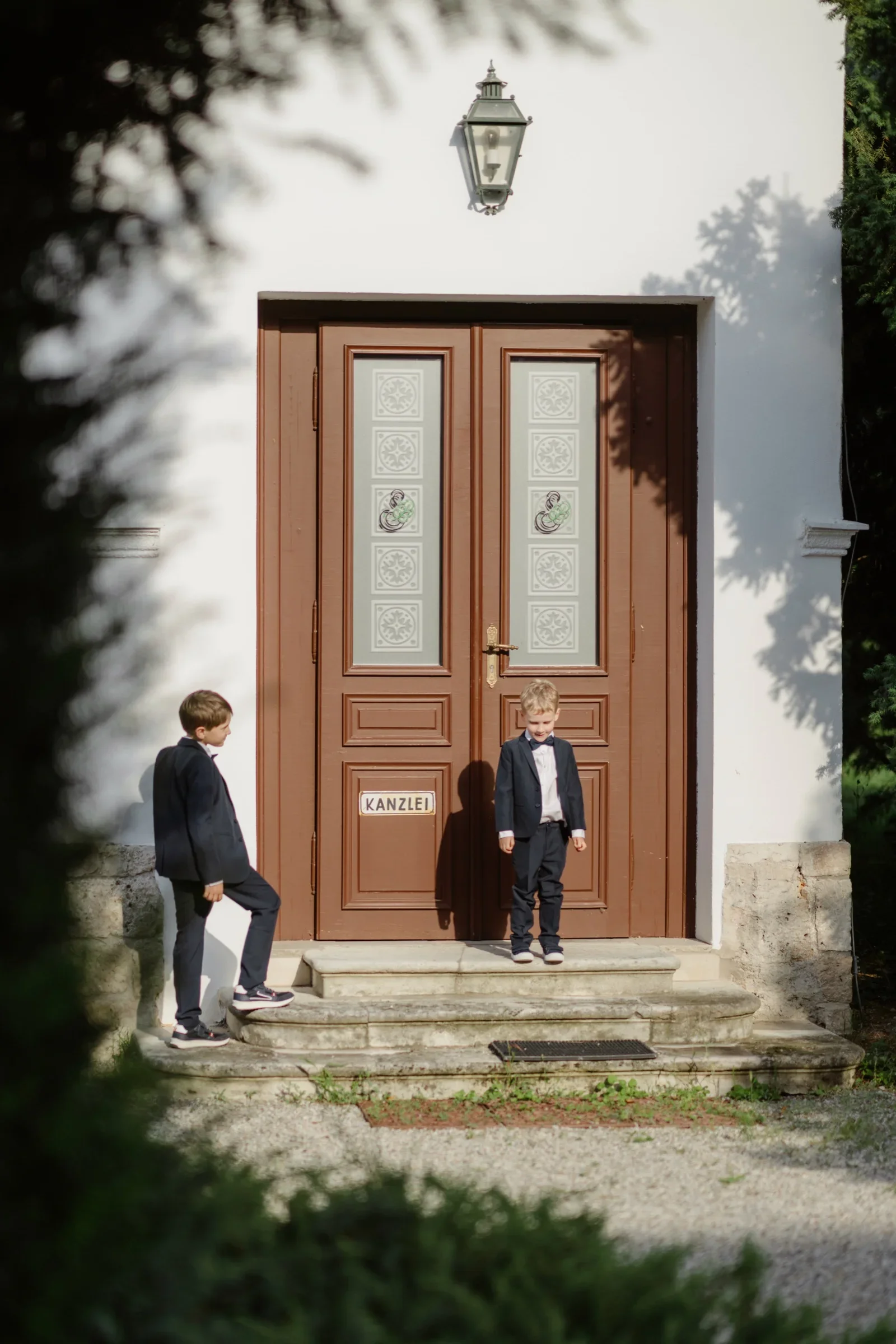 Two boys in dark suits sitting at a wooden double door of the villa