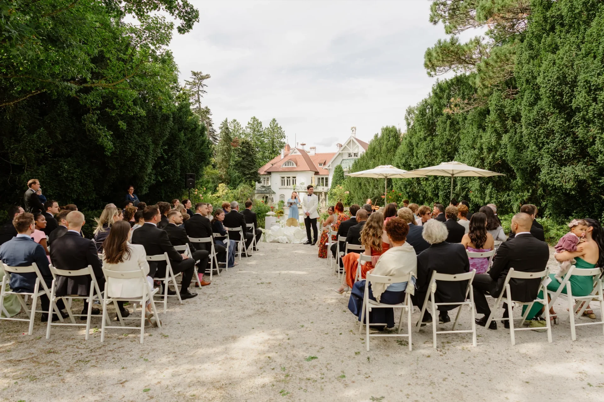 Guests seated on white chairs for an outdoor ceremony under tall trees