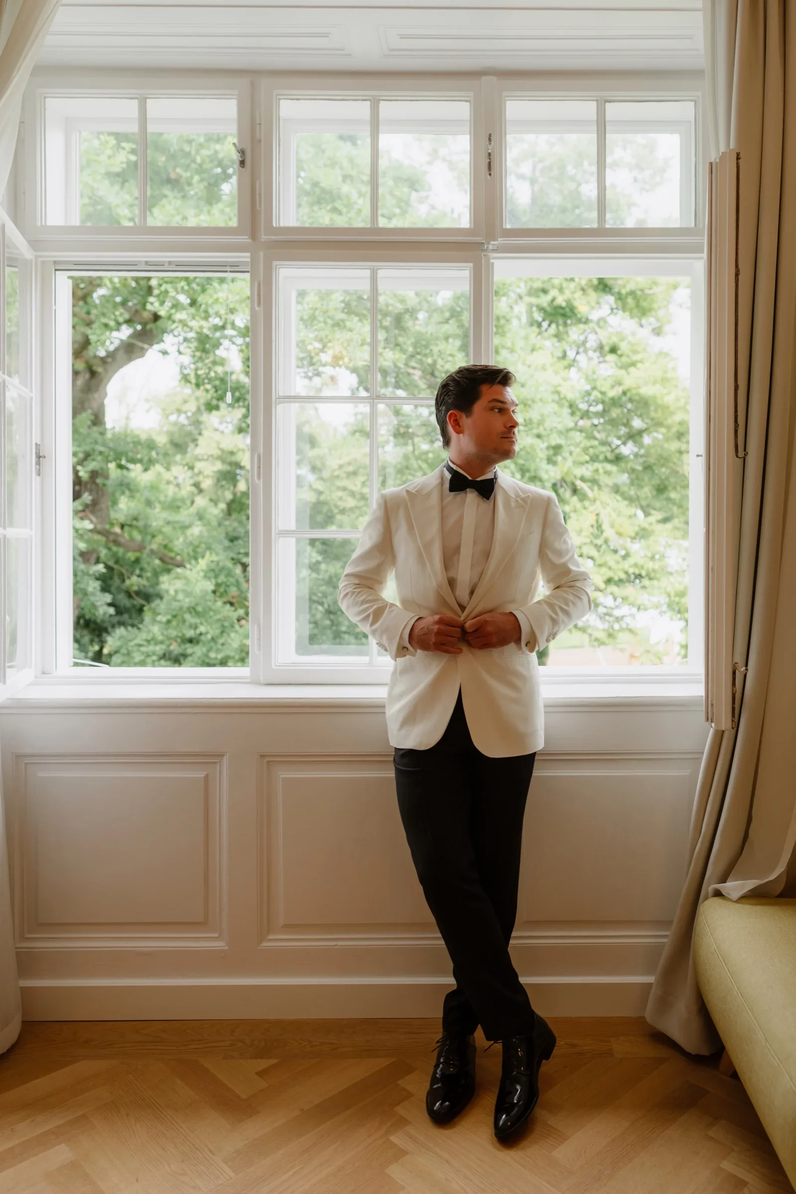 Groom in a white dinner jacket standing with crossed legs in front of tall windows to the park