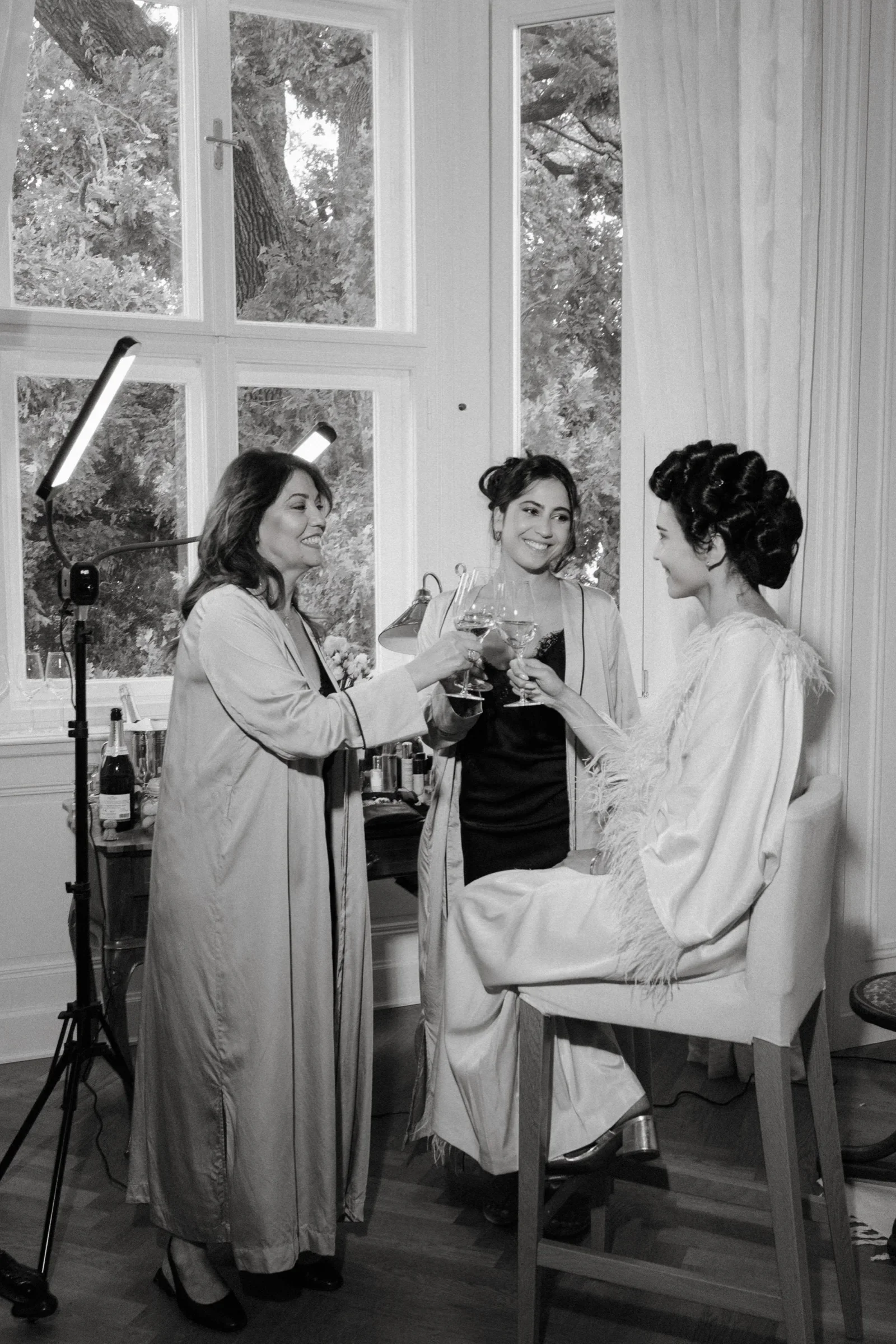 Bride getting ready with helpers beside a window and a light stand