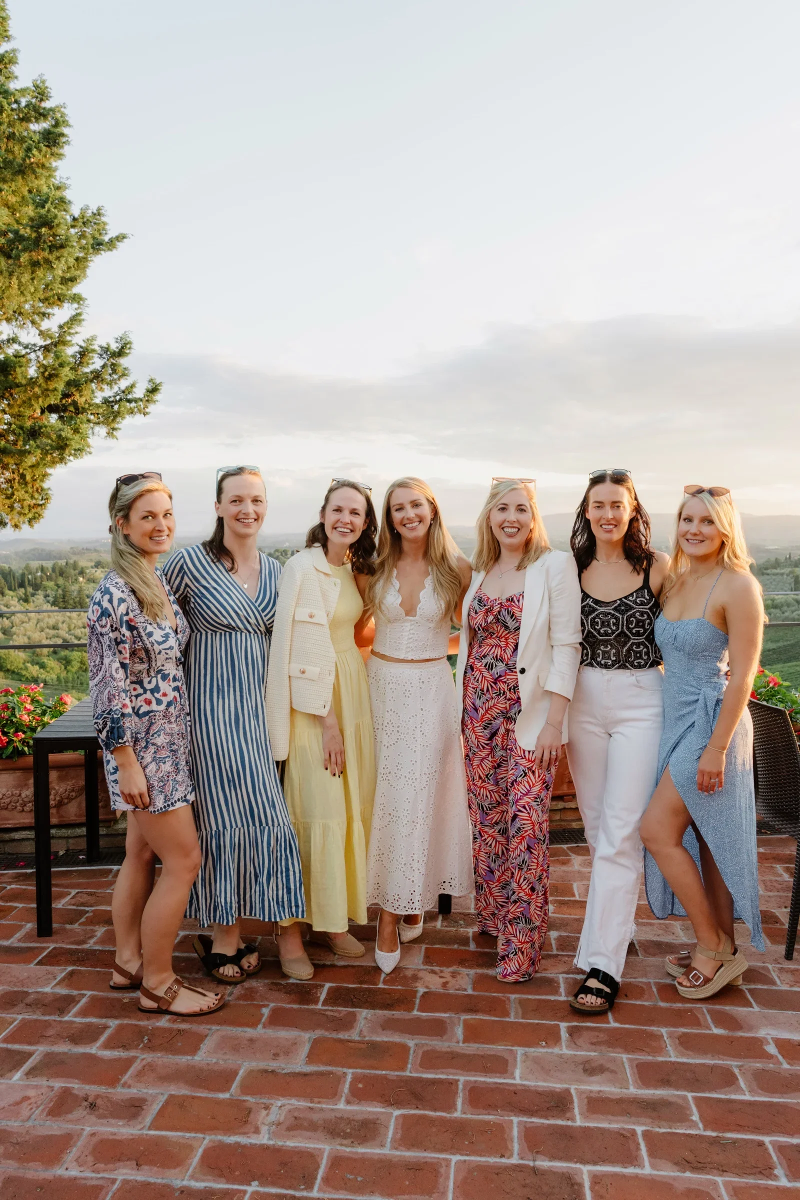 Seven women in summer dresses laughing together on the villa's brick terrace at sunset