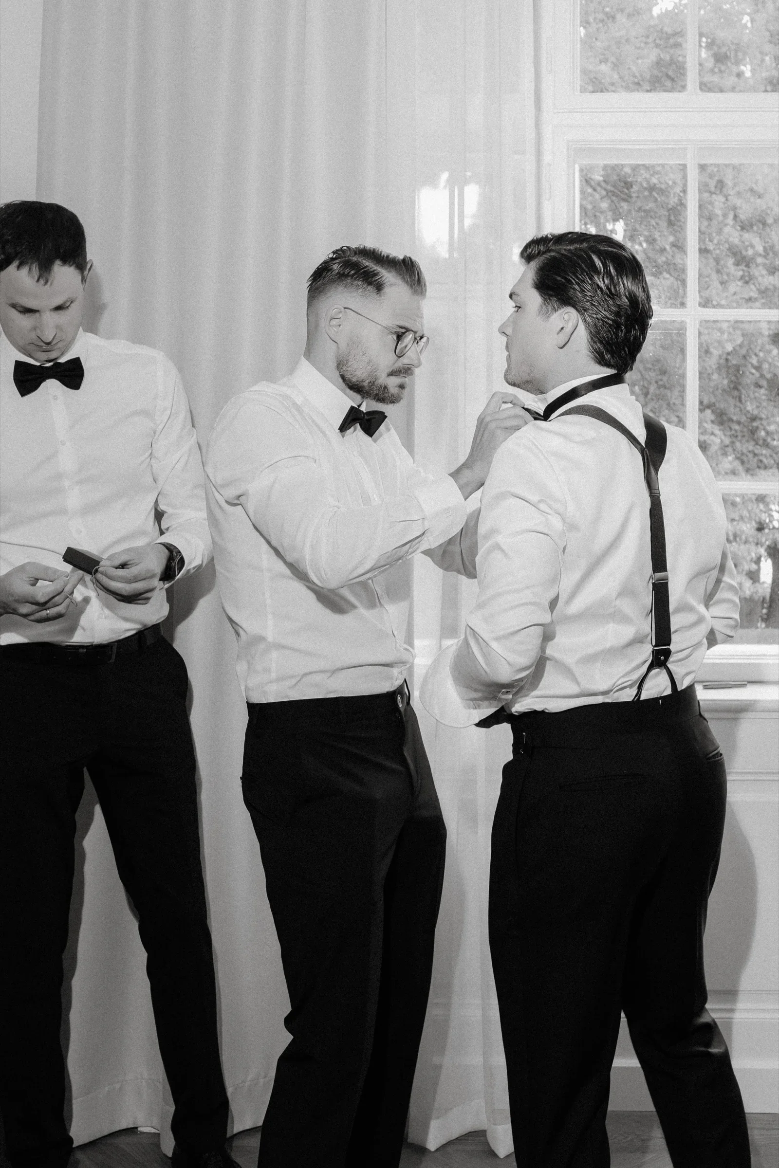 Black and white photo of three groomsmen in bow ties and suspenders getting ready by a window