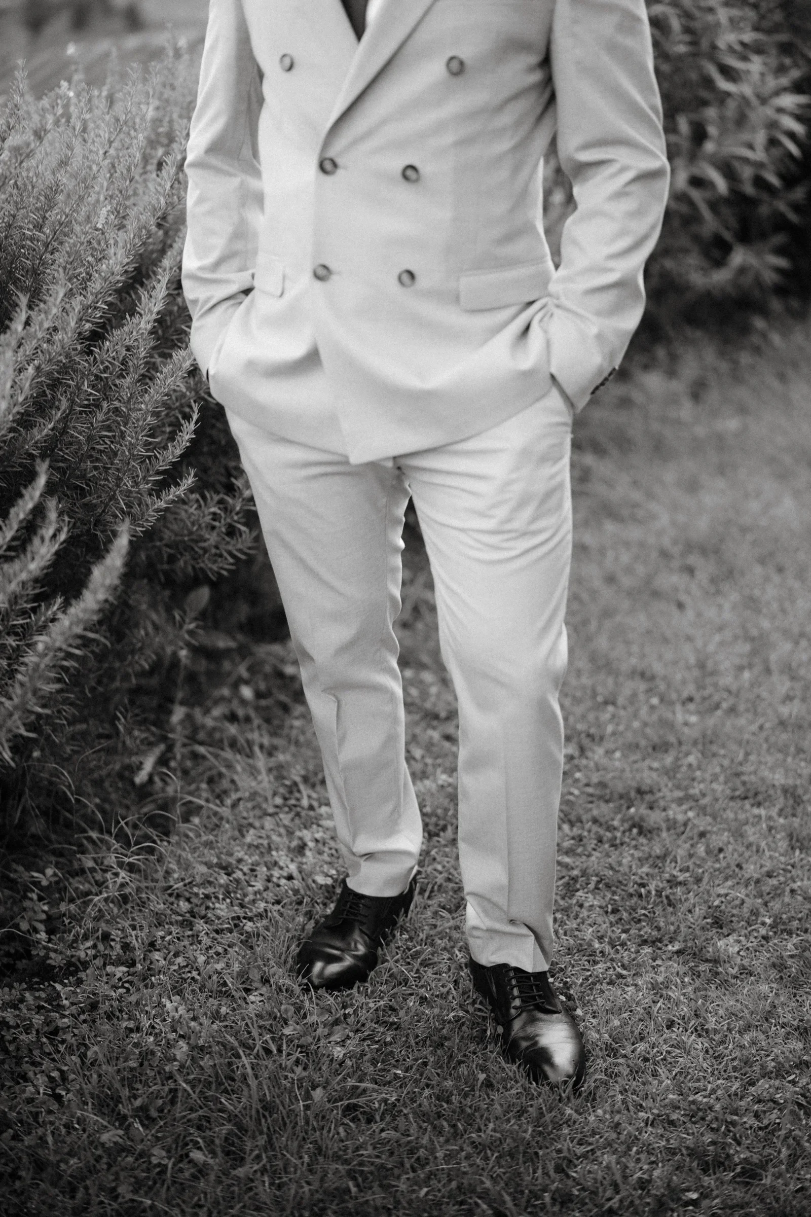 Black and white: the groom standing in long grass, hands in his pockets