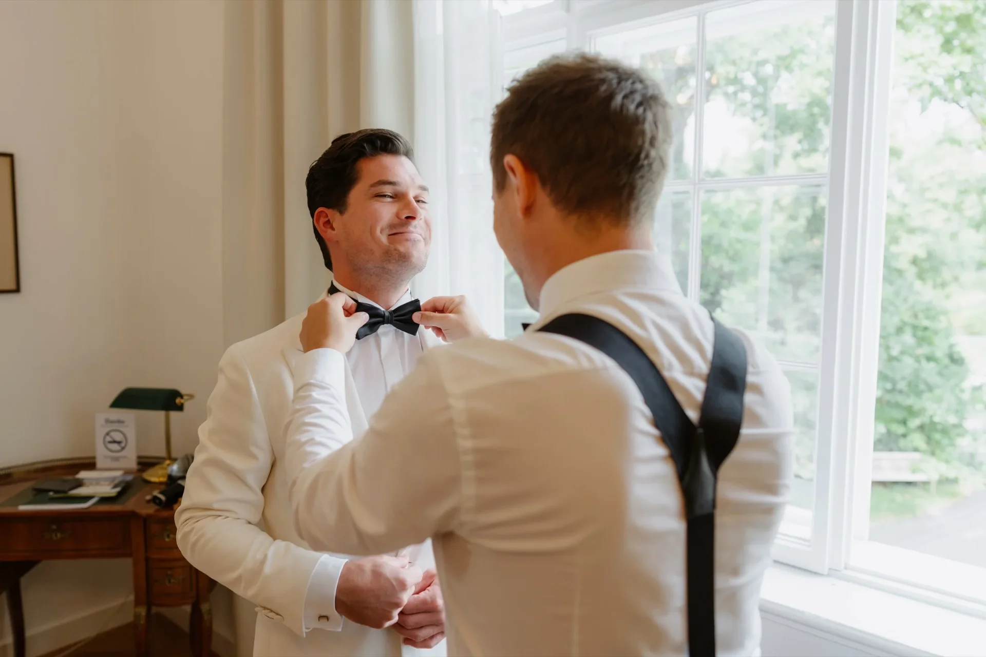 Groomsman tying the groom's black bow tie by a sunlit window while the groom smiles