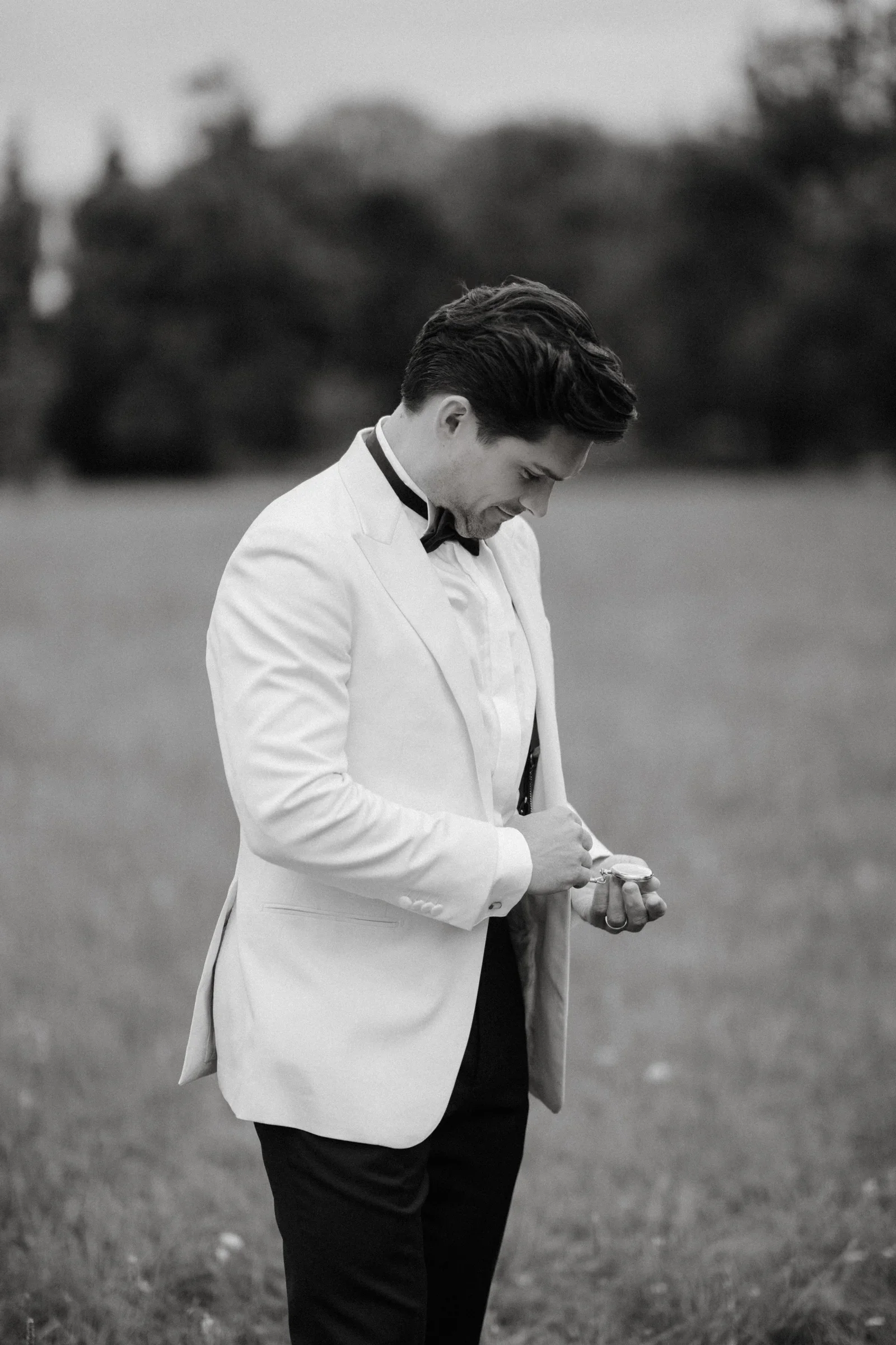 Groom in a white jacket checking his cuff in a meadow, black and white