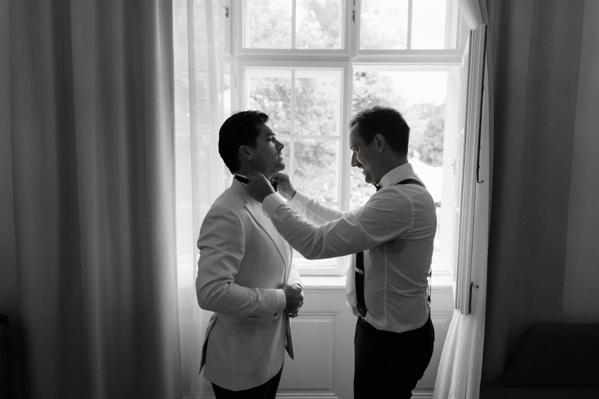 Black and white photo of a groomsman fixing the groom's collar beside a tall window