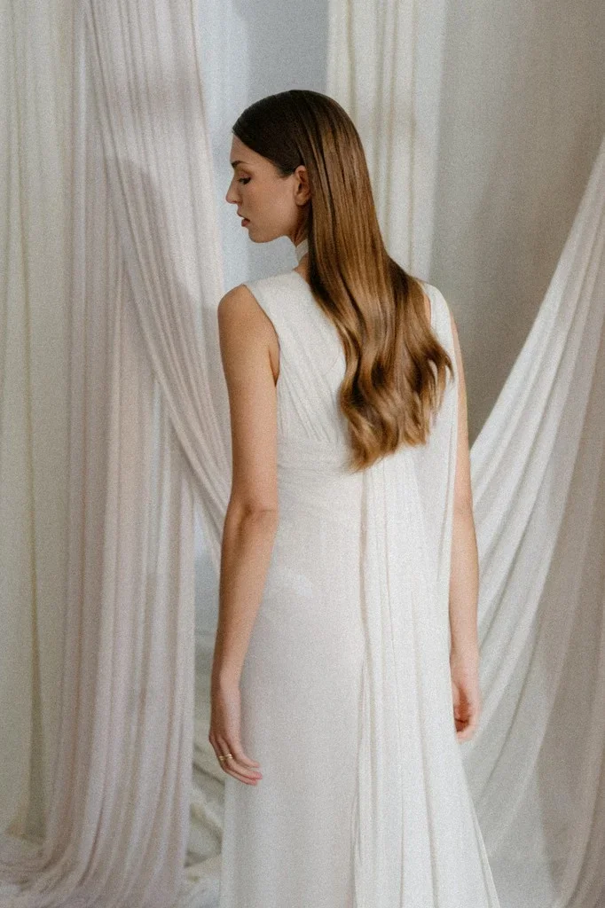 Bride seen from behind in a column gown before pale drapes