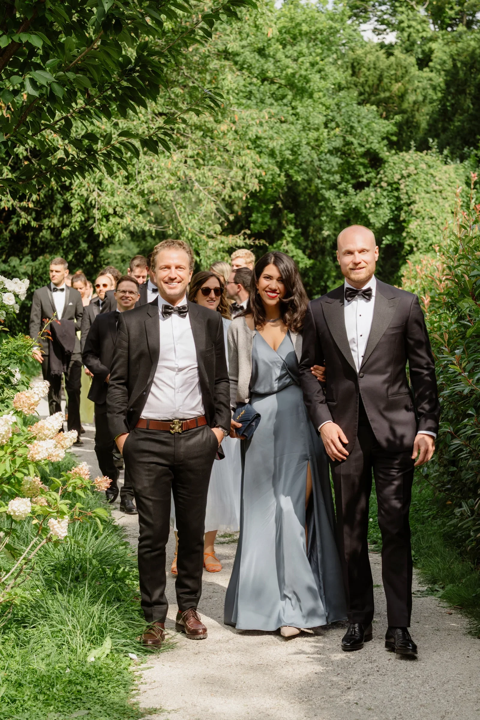 Groom and two companions walking down a garden path lined with roses