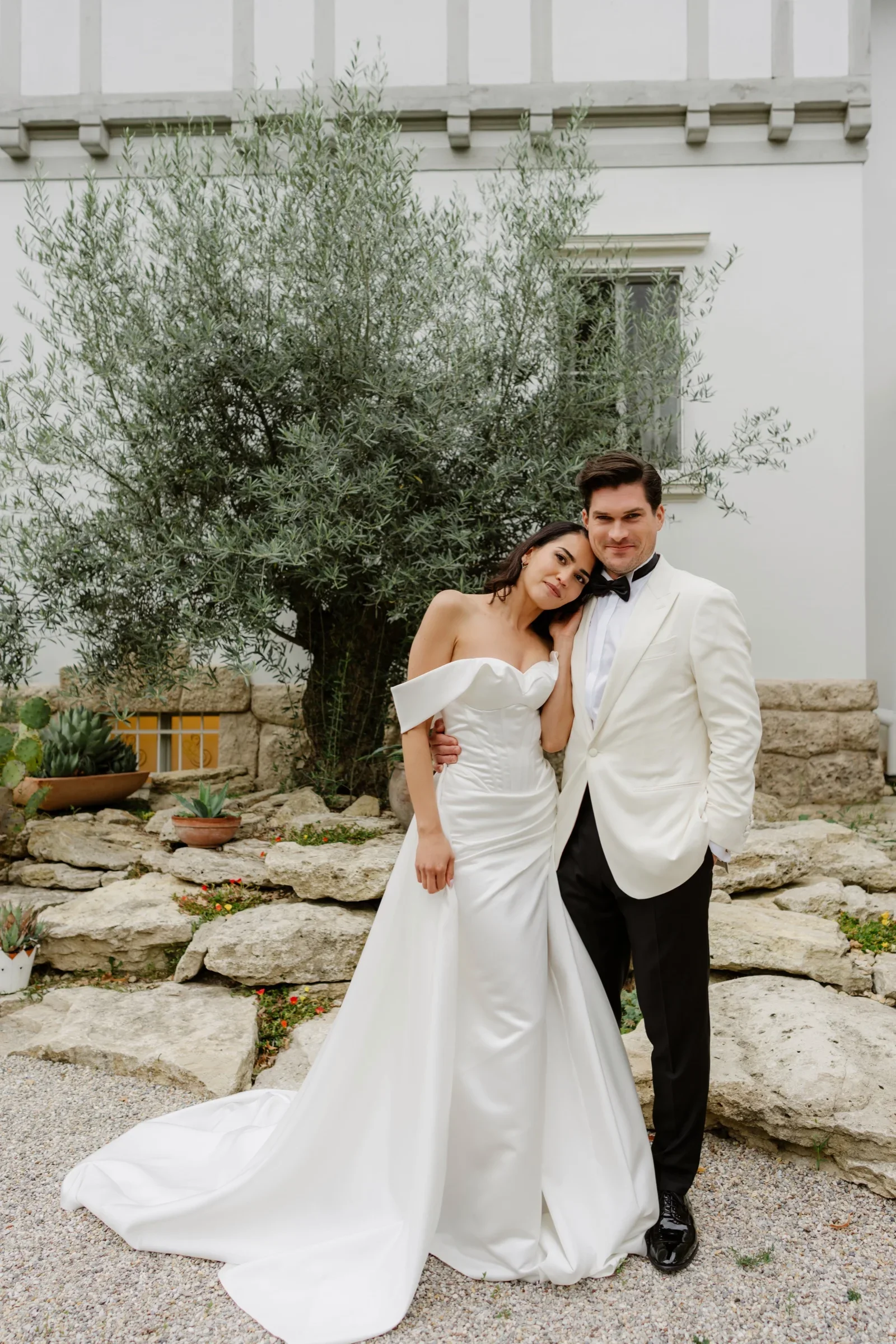 Couple posing in front of a white villa with an olive tree, the bride's train spread on the gravel