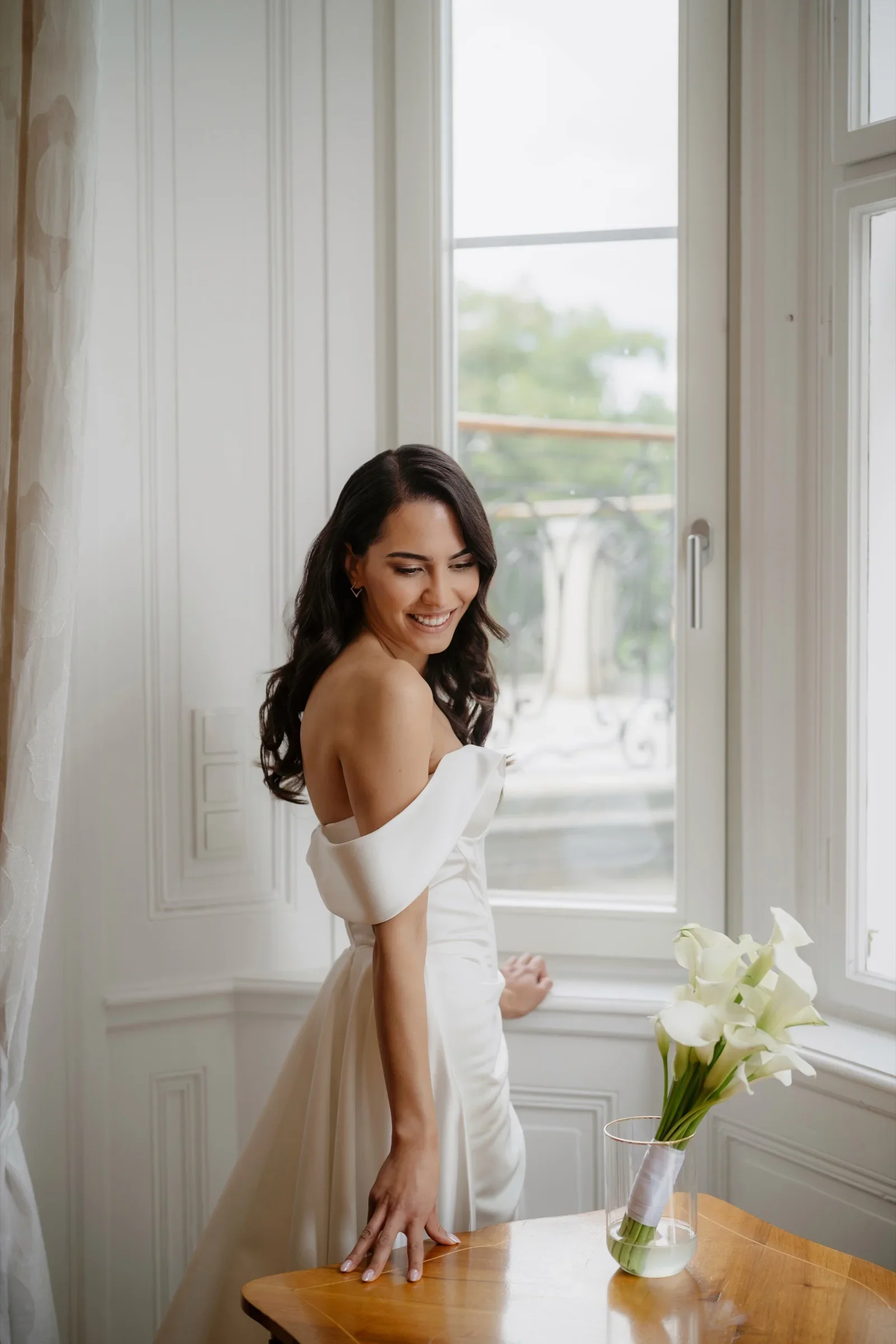Smiling bride in a white dress by a window leaning on a table with a small bouquet