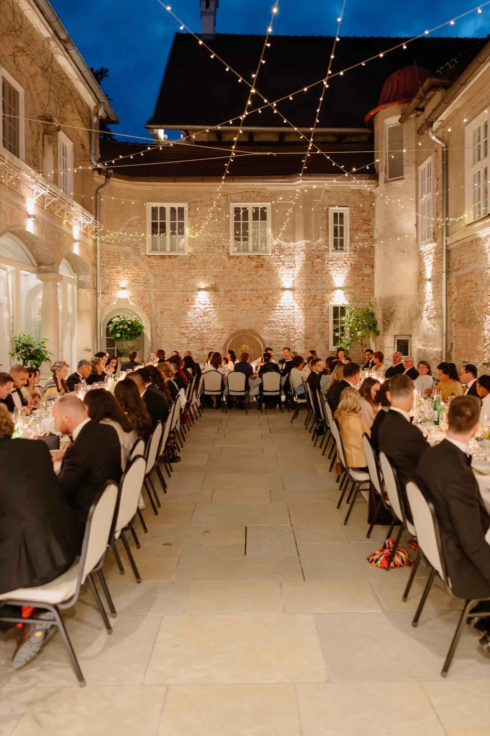 Guests seated at long tables in the castle courtyard under string lights at blue hour