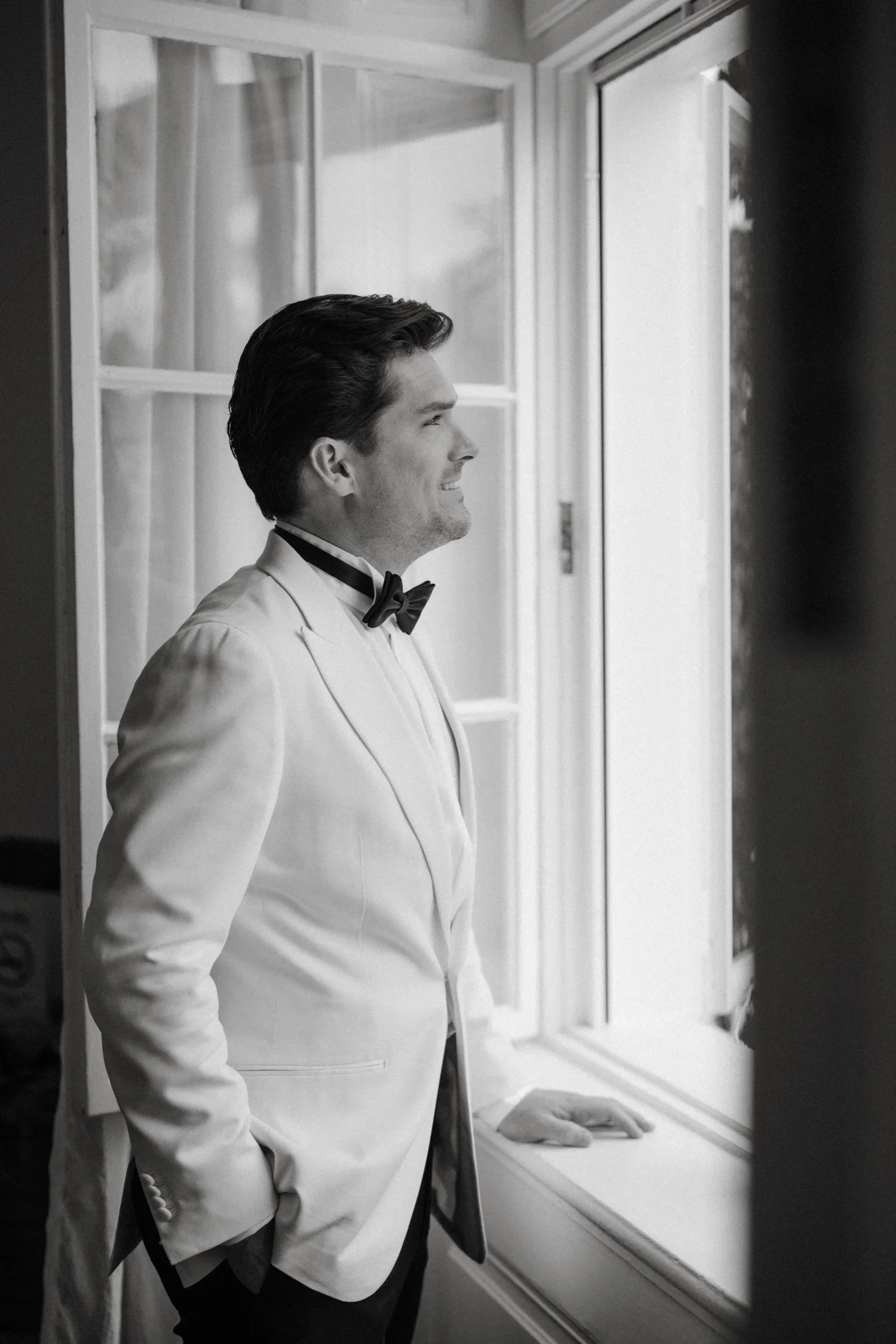 Black and white photo of the groom in a white dinner jacket leaning at a window