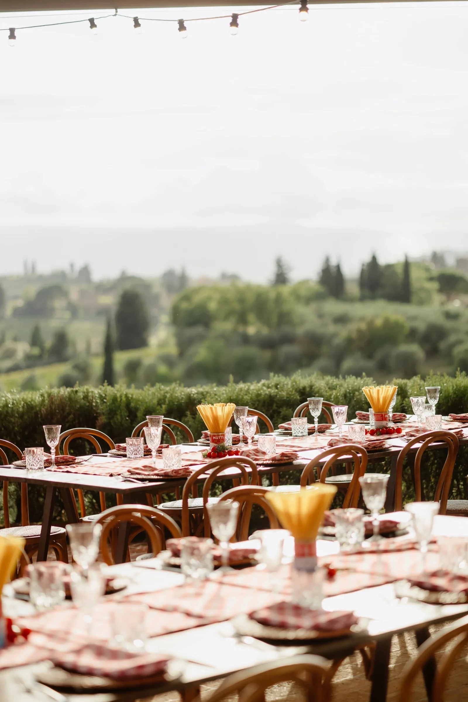 Long welcome-dinner tables under festoon lights, set with red gingham and looking out over the Tuscan hills