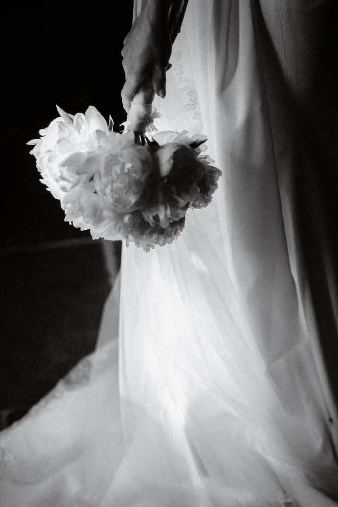 Hand holding white peonies against a pale gown in low light