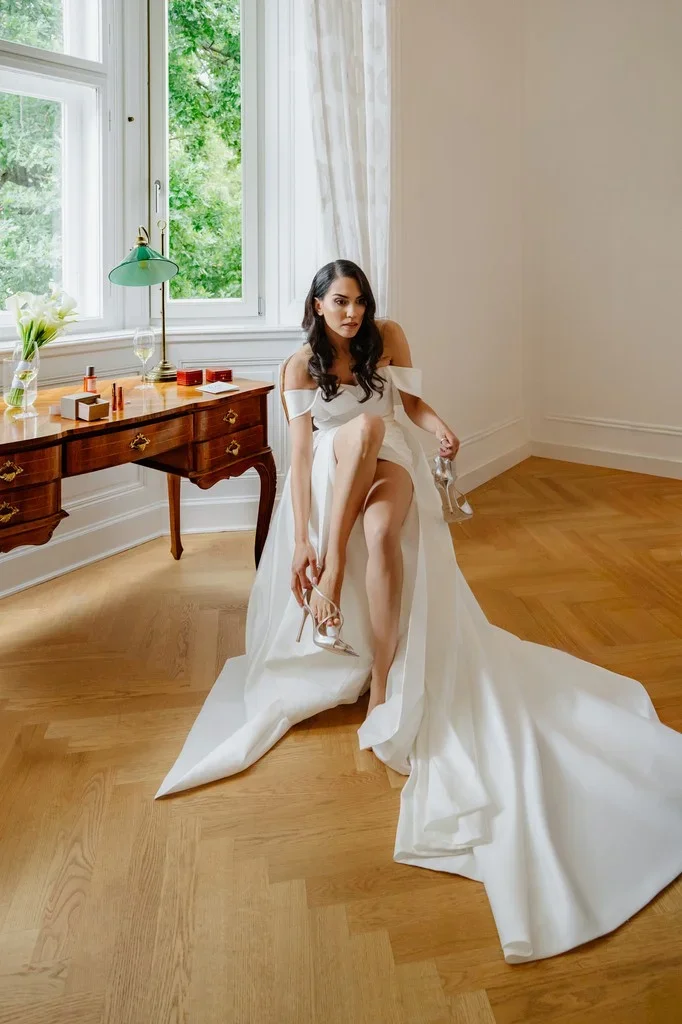 Bride fastening her shoe on a parquet floor beside a sunlit window