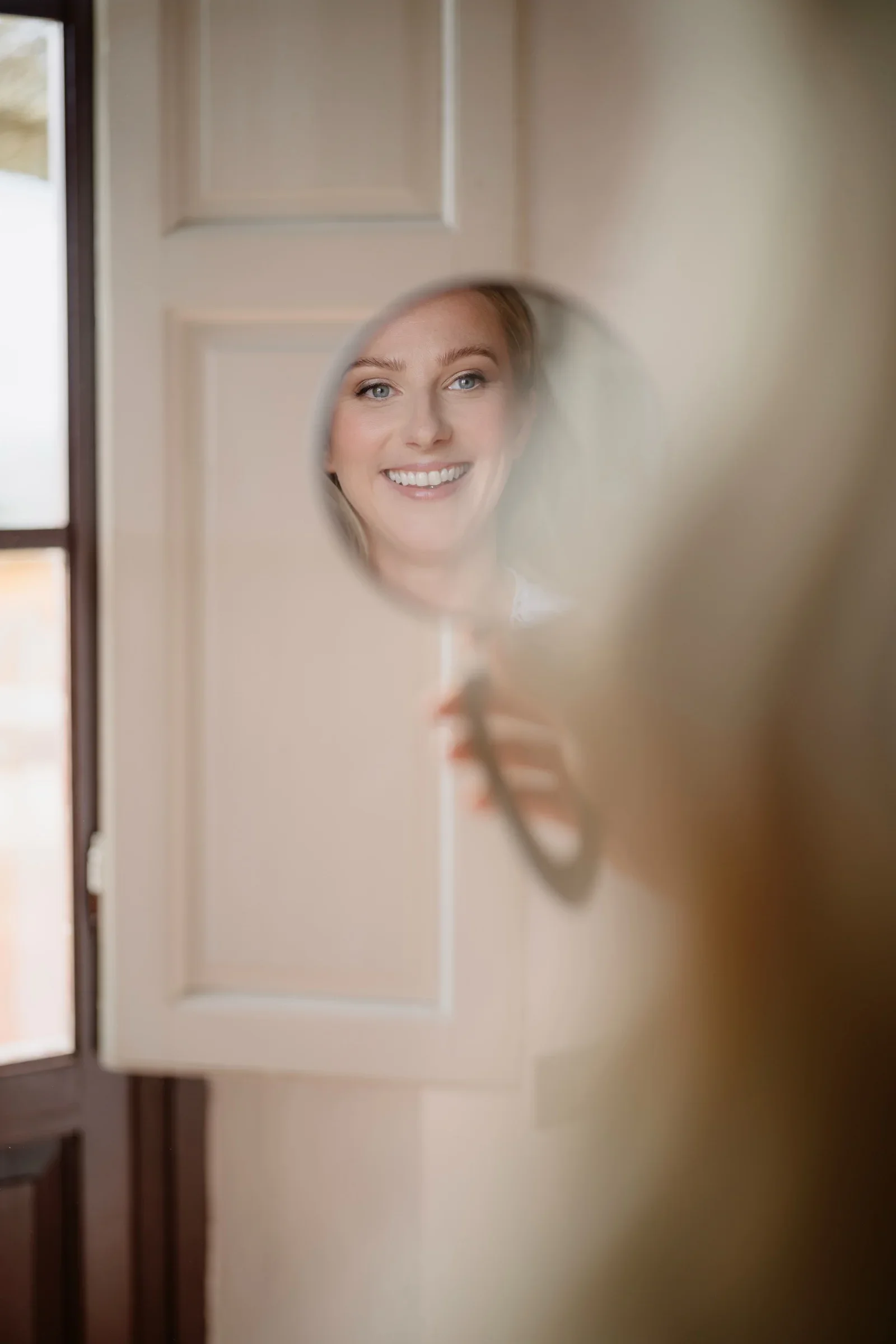 The bride's face reflected in a round hand mirror as she smiles