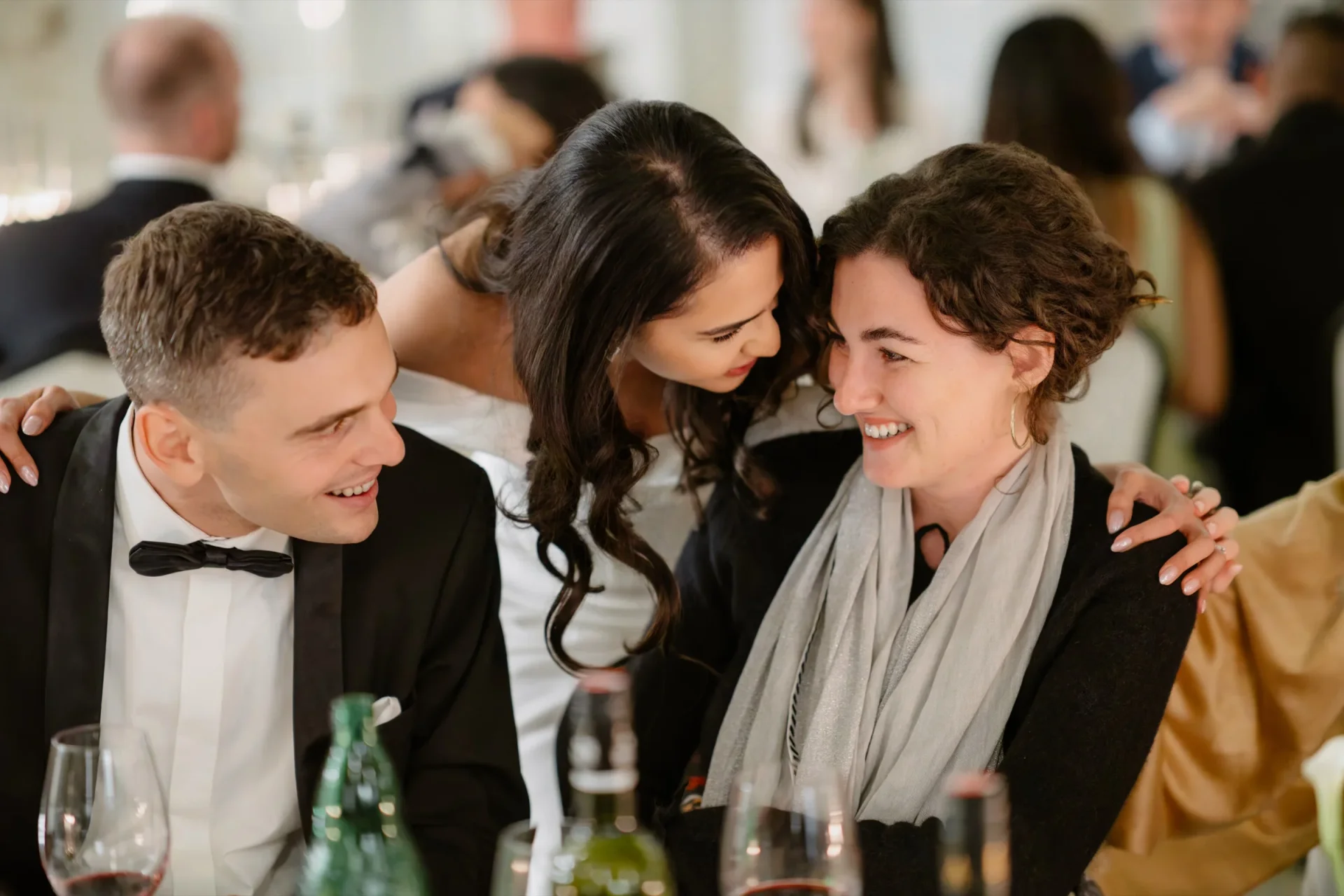 Groom, bride and a woman in a gray scarf laughing together at the table